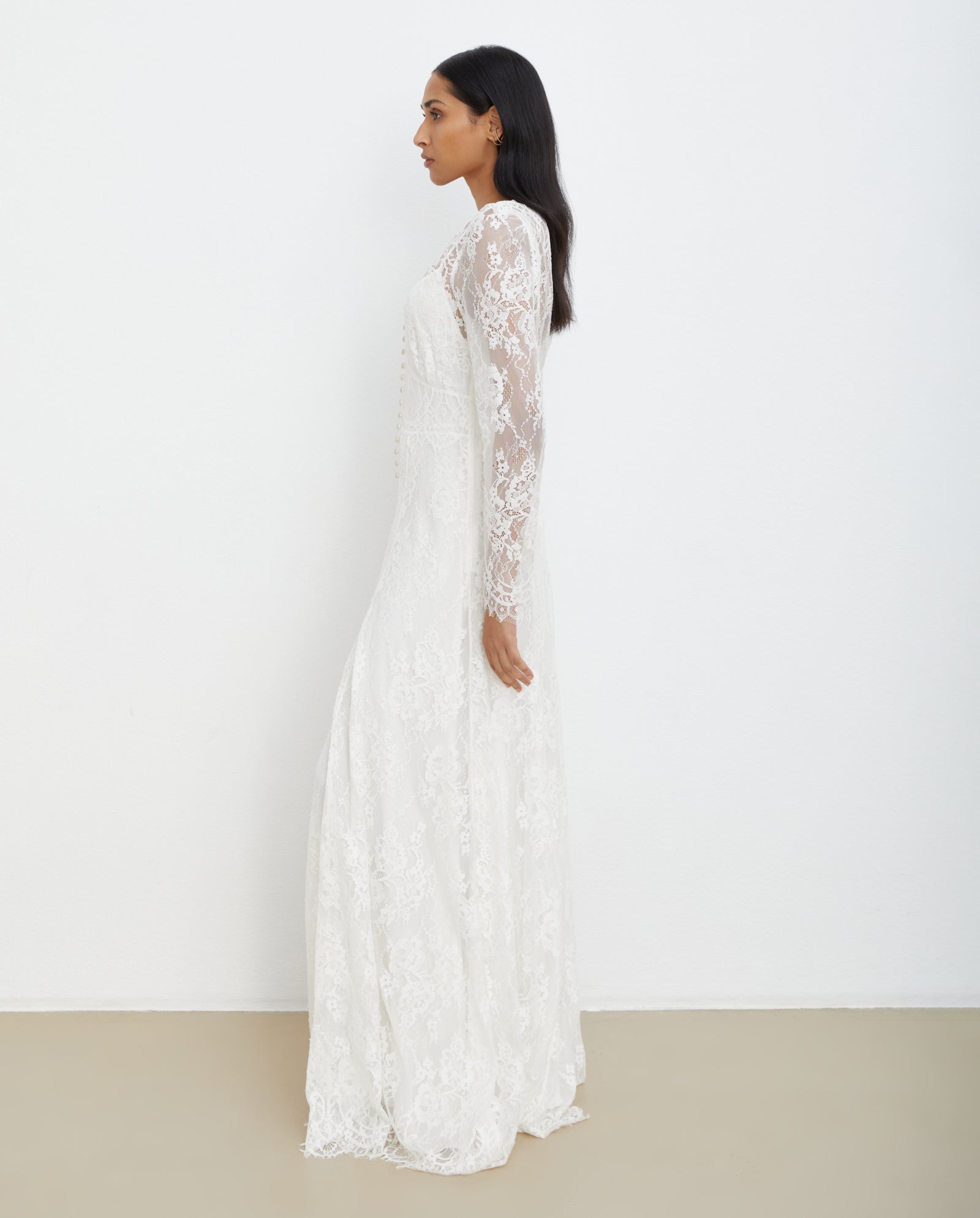 A woman wearing the IVY OAK MARCITA Bridaldress—a long-sleeved, lace white maxi wedding dress with a deep V-neckline—stands sideways against a plain white wall.