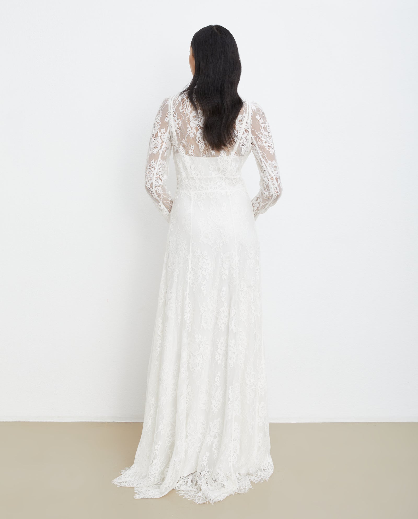A woman stands with her back to the camera wearing the IVY OAK MARCITA Bridaldress, a long-sleeved, floor-length white lace gown with a scalloped hem.