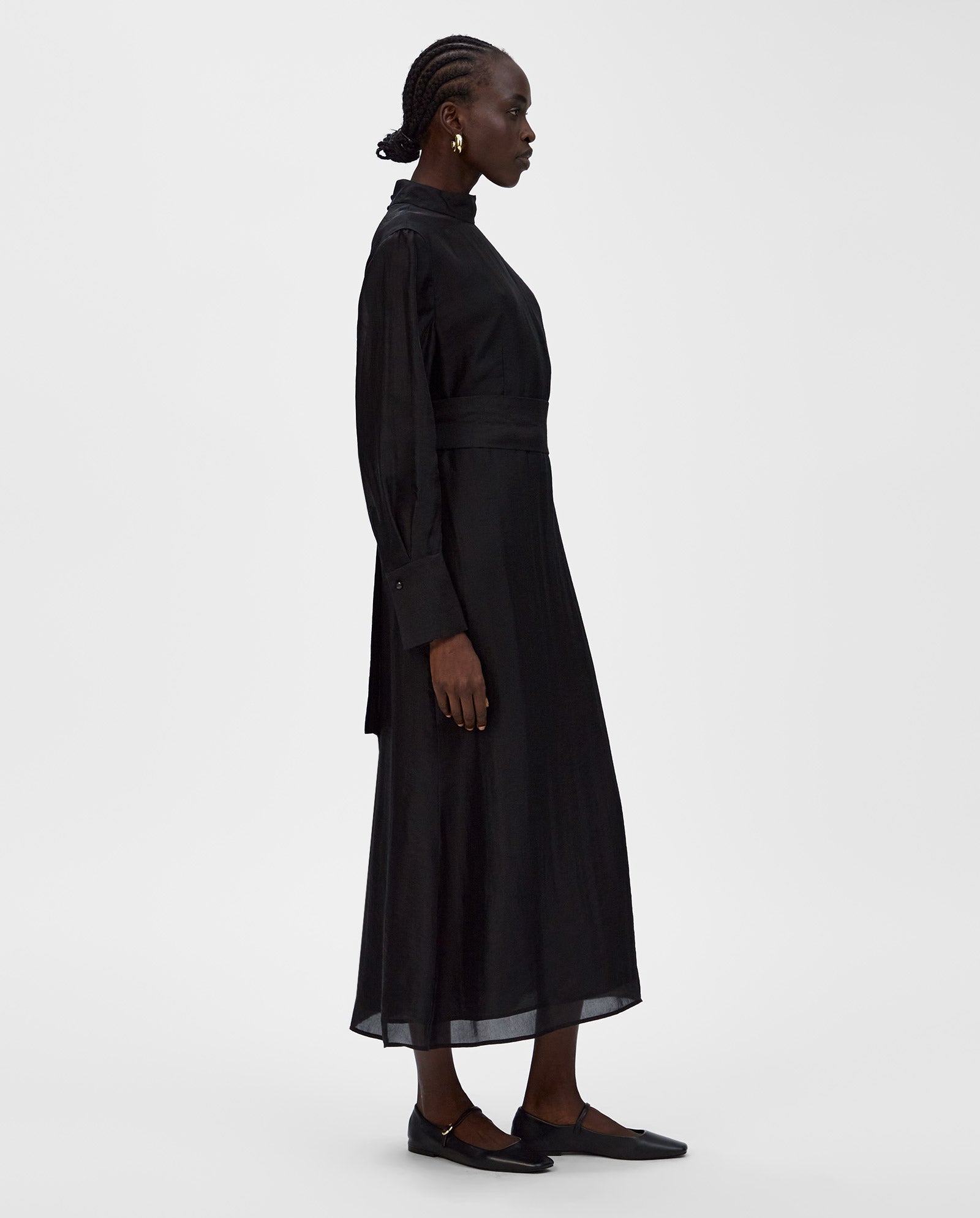 Woman in the MALINA long-sleeved, high-neck midi black dress by IVY OAK with black flats, standing sideways against a white background.