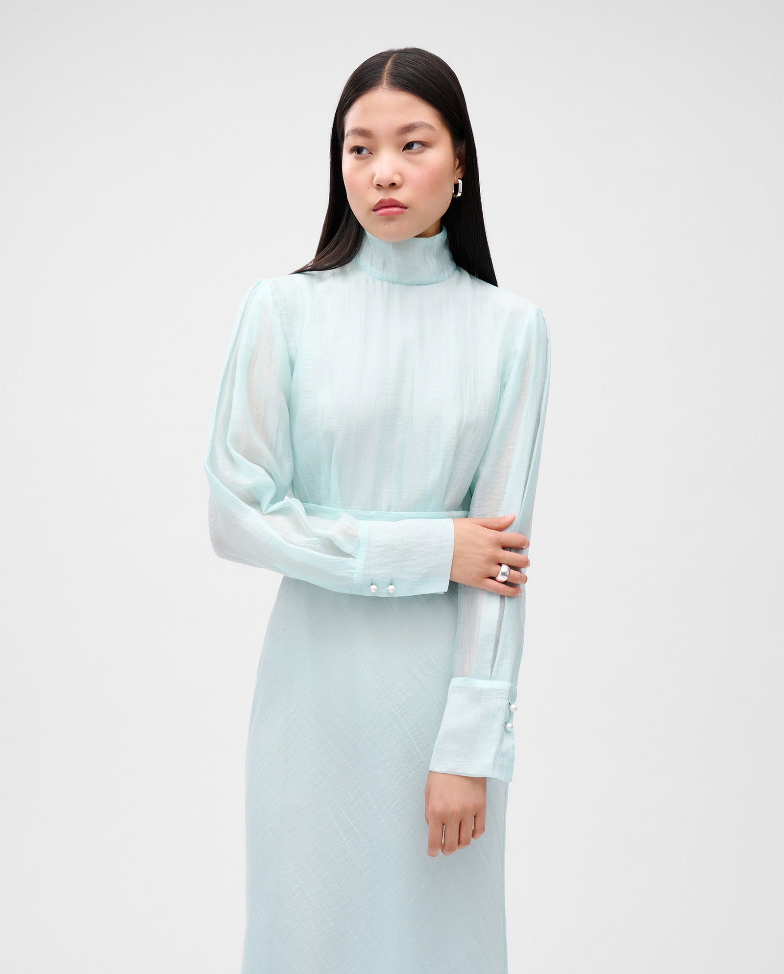A woman wears the IVY OAK MALINA dress—light blue, midi-length with long sleeves, a high neck, and an elegant flared skirt—standing against a plain light gray background.