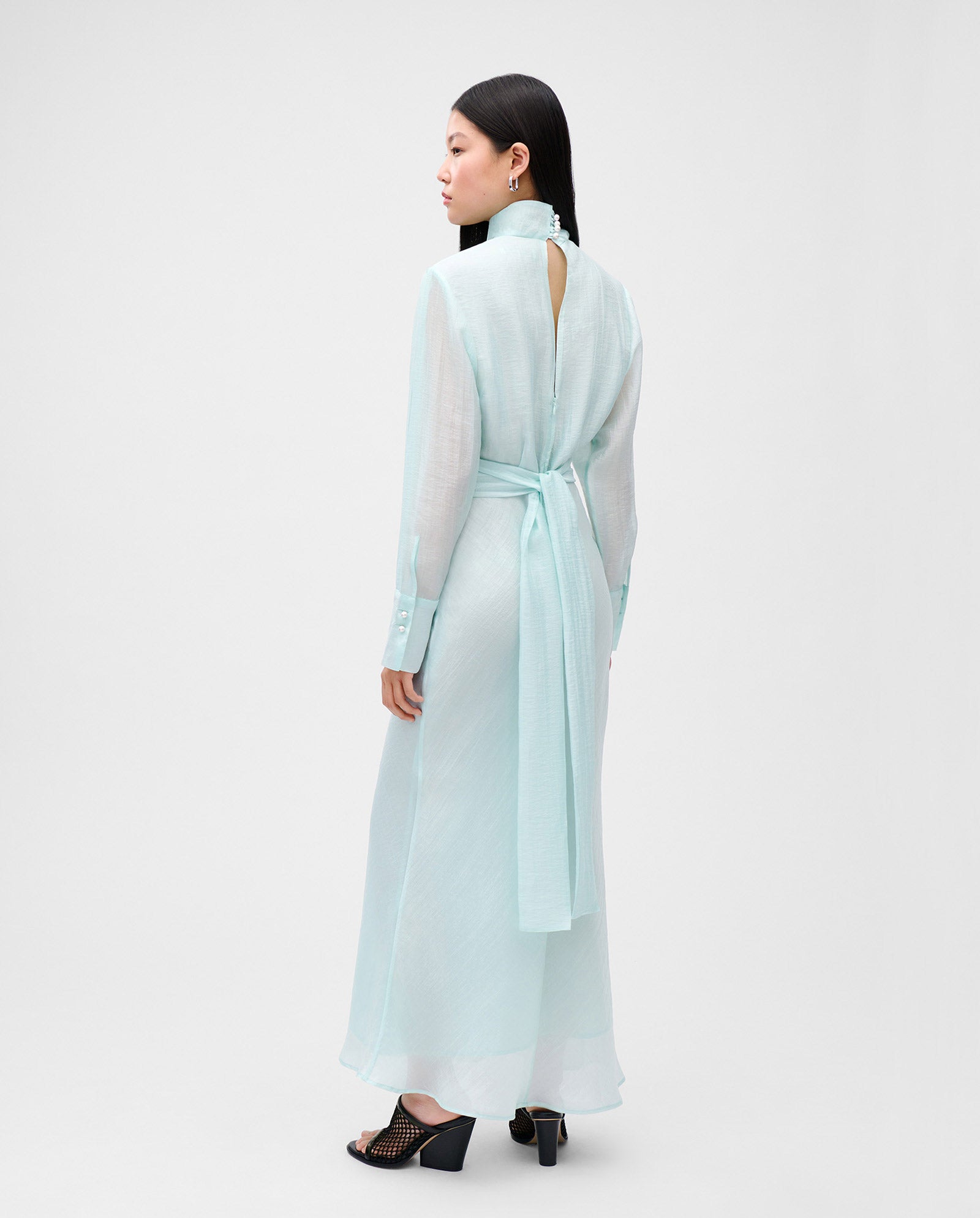 A woman stands facing away in a light blue IVY OAK MALINA midi dress featuring long sleeves, a belt, and a flared skirt.
