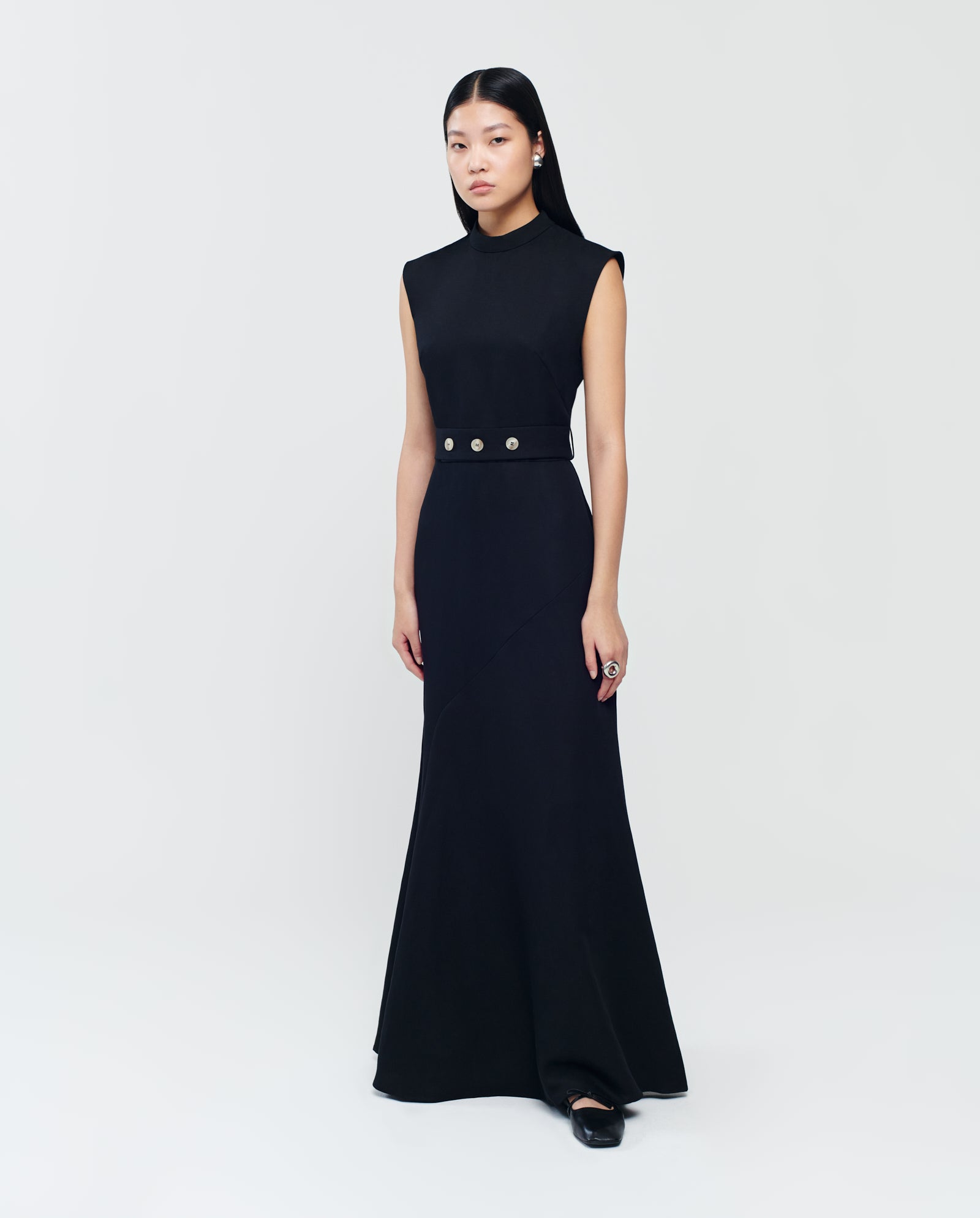 A woman wears the IVY OAK MANILA Dress—a sleeveless maxi with a belted waist and stand-up collar—standing against a plain light background.