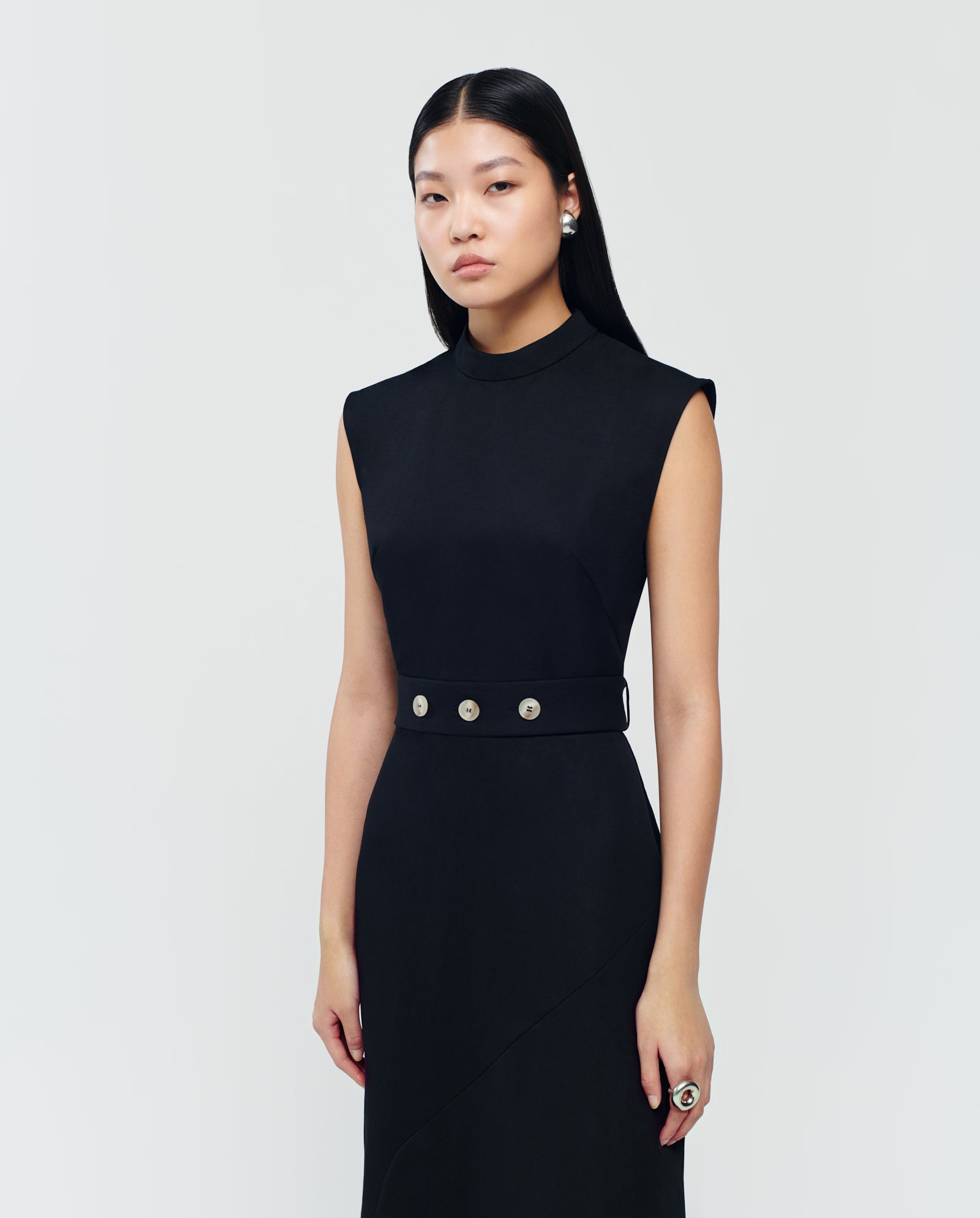 A woman with long black hair wears the IVY OAK MANILA Dress, a sleeveless maxi dress featuring silver buttons and a stand-up collar, standing against a plain background.