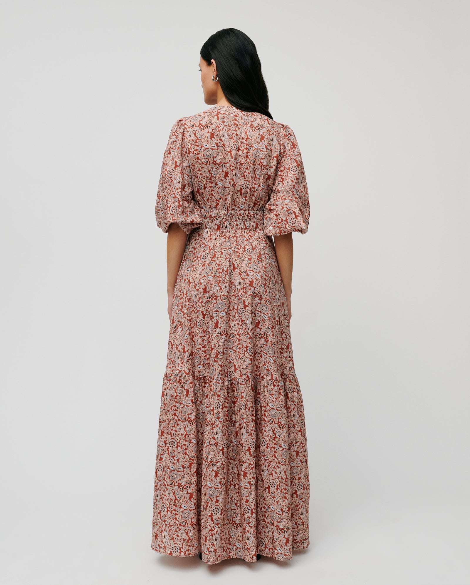 Woman standing with her back to the camera, wearing the IVY OAK DREA Dress—a long floral V-neck maxi with puffed sleeves.