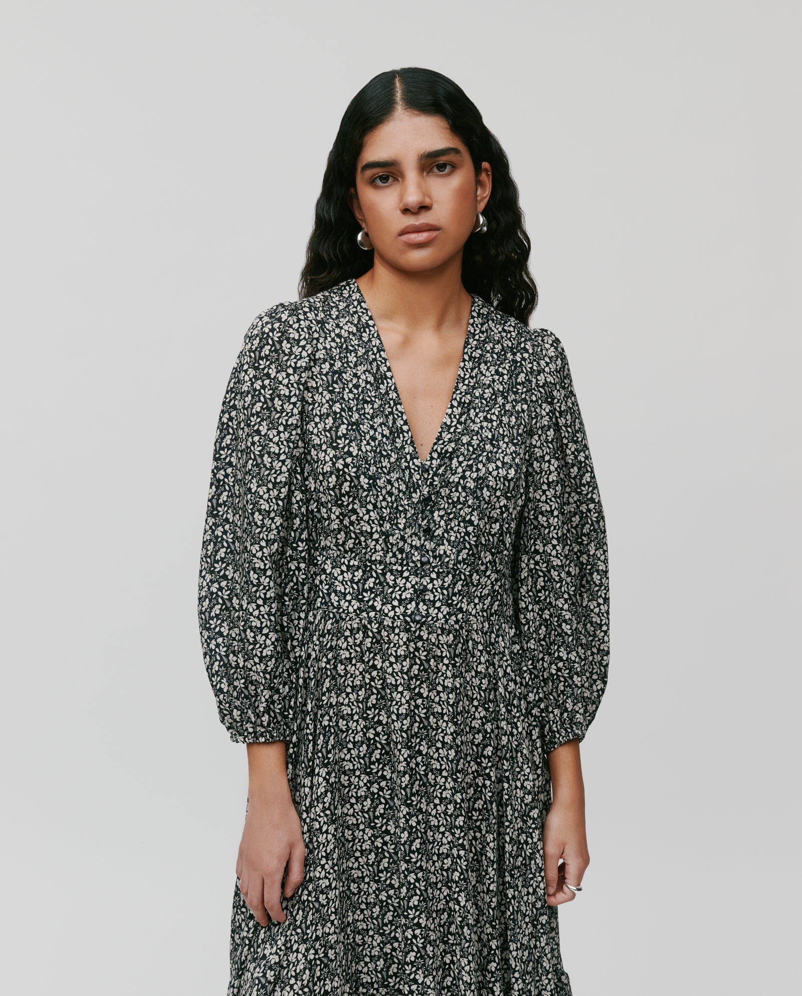 A woman with long wavy dark hair wears the IVY OAK DREA Dress, featuring a black floral print, balloon sleeves, and a V-neck, set against a plain background.