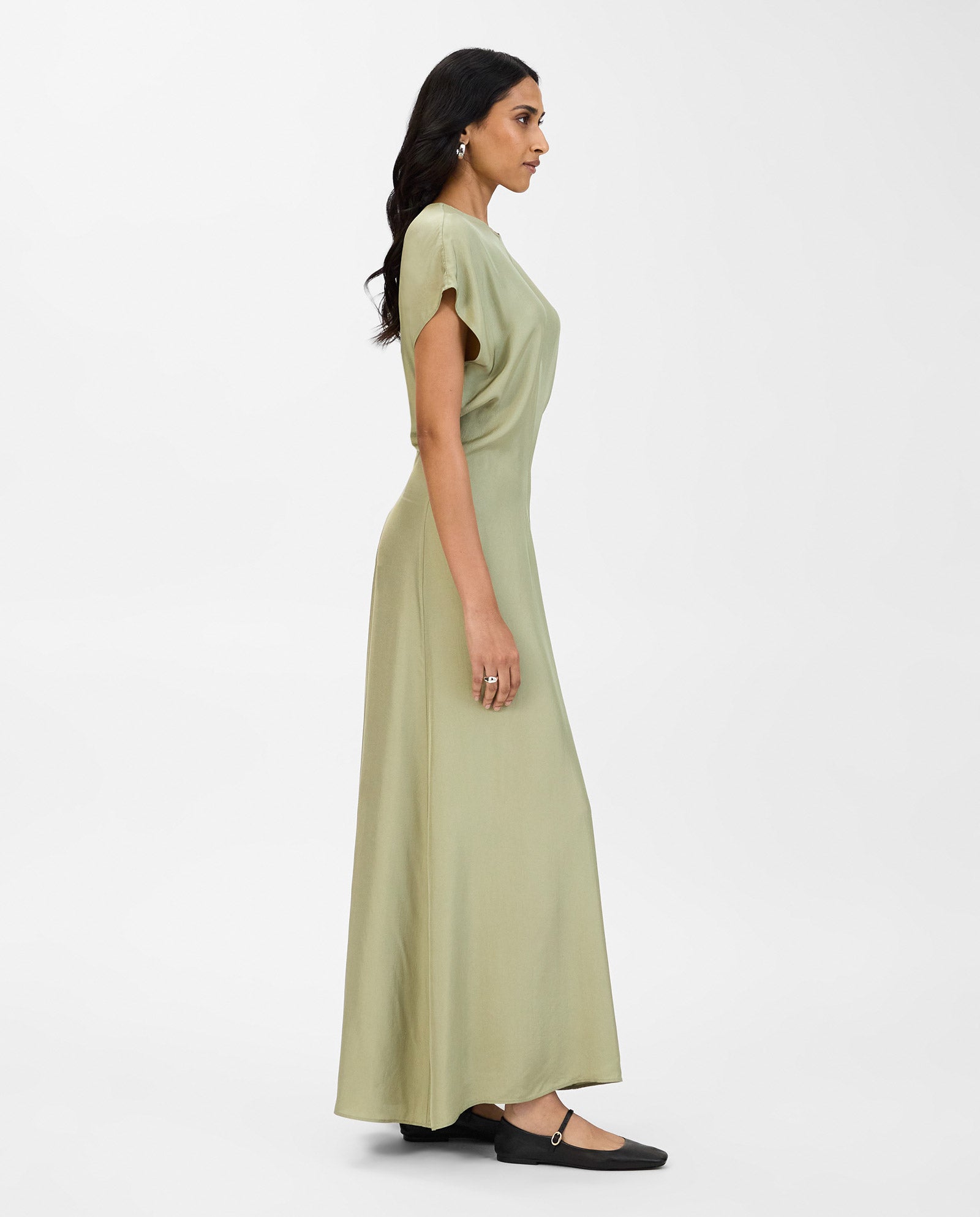A woman in the IVY OAK MARLIS SUE dress—sage green, fitted waist, short sleeves, NAIA acetate—paired with black flats, stands in profile against a white background.