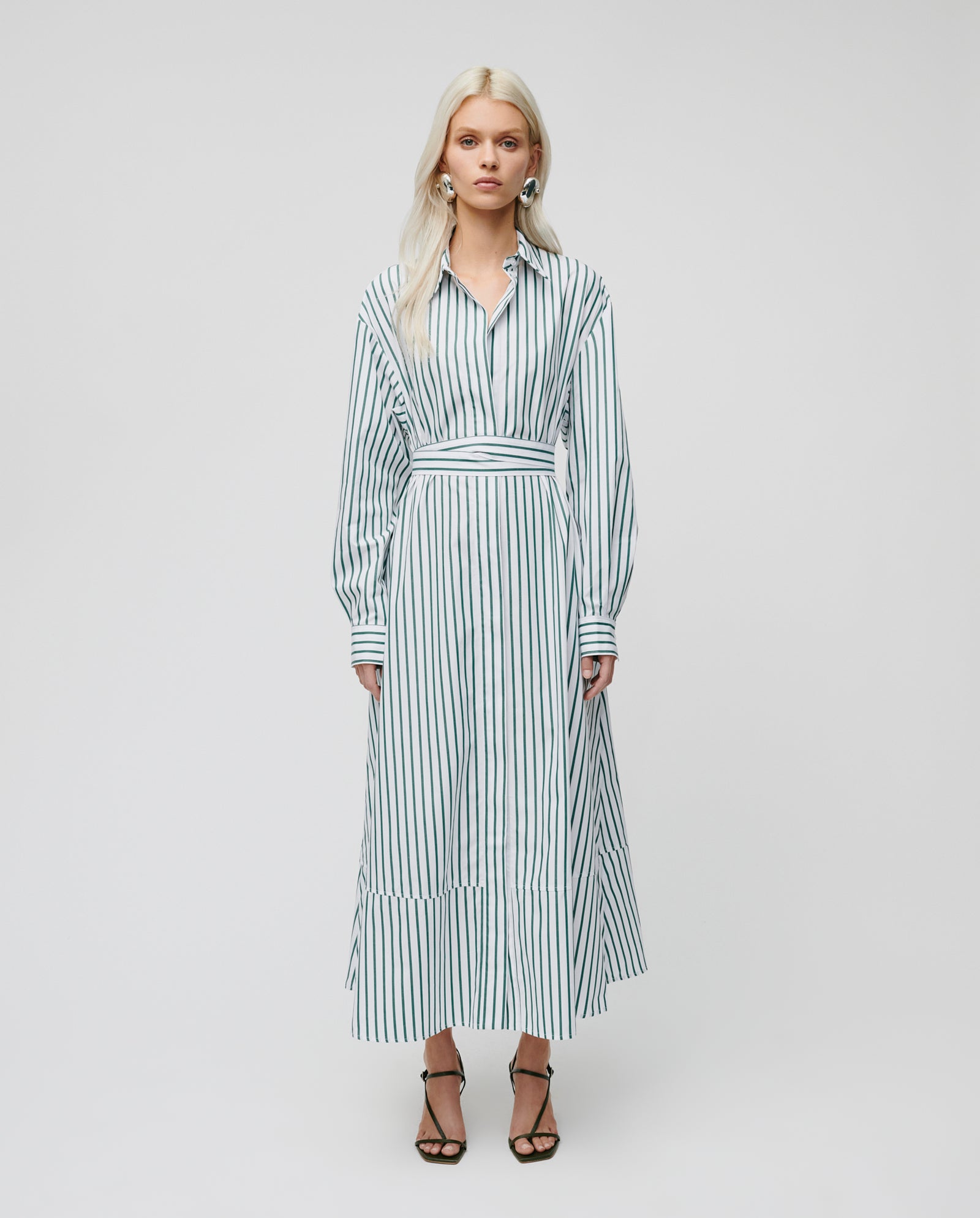 A woman stands facing forward in black strappy heels, wearing IVY OAK’s DINA KIM Dress—a long-sleeved white dress with vertical blue stripes, organic cotton fabric, and side pockets.