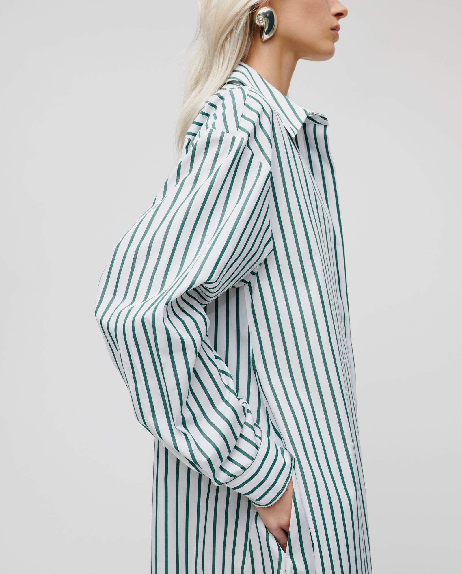 Woman in a green and white striped DINA KIM dress by IVY OAK, made from organic cotton, hand in side pocket, wearing statement earrings.