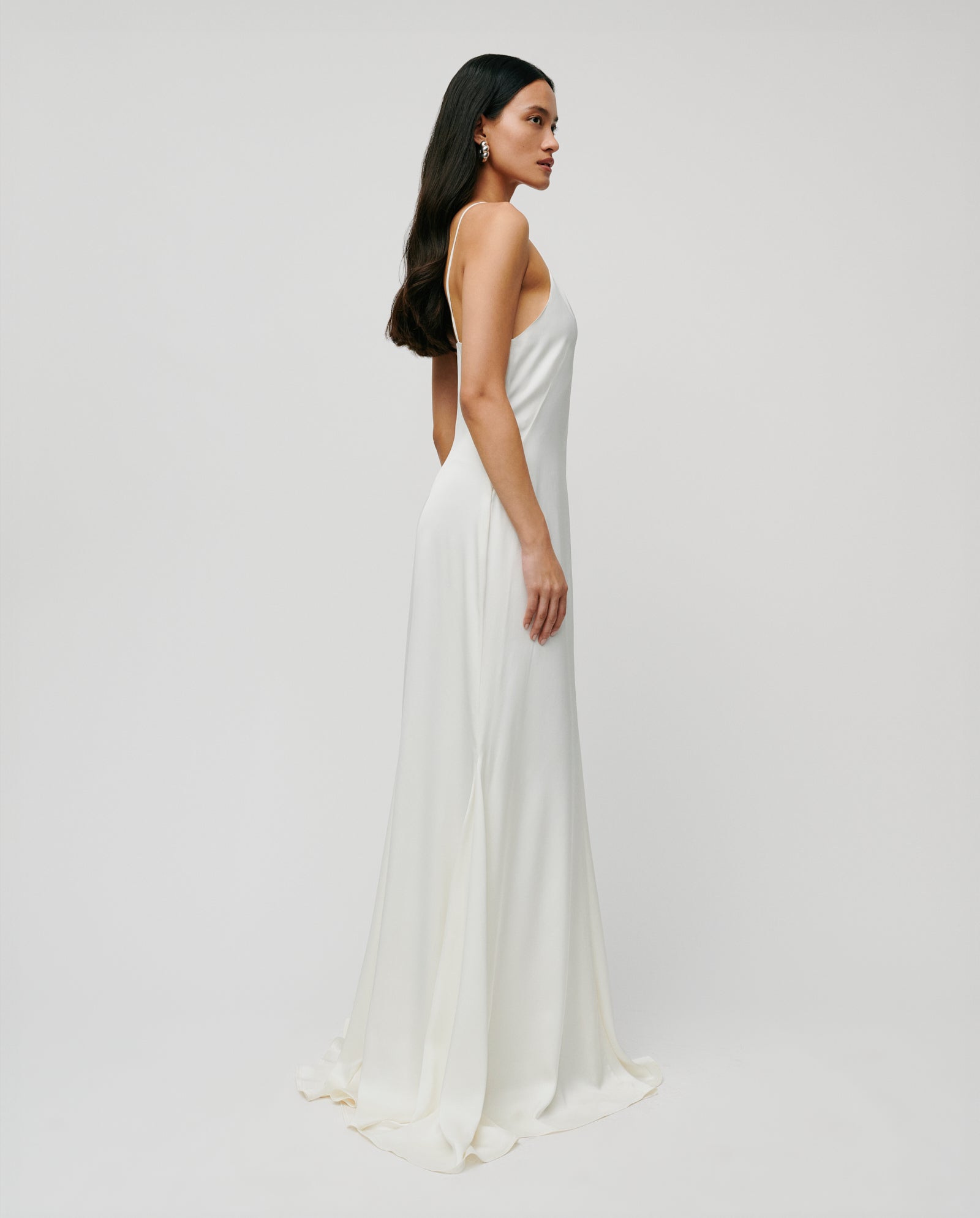 A woman stands sideways against a plain, light background wearing the IVY OAK MADELIN IDA Bridaldress—a long, white, sleeveless maxi gown.