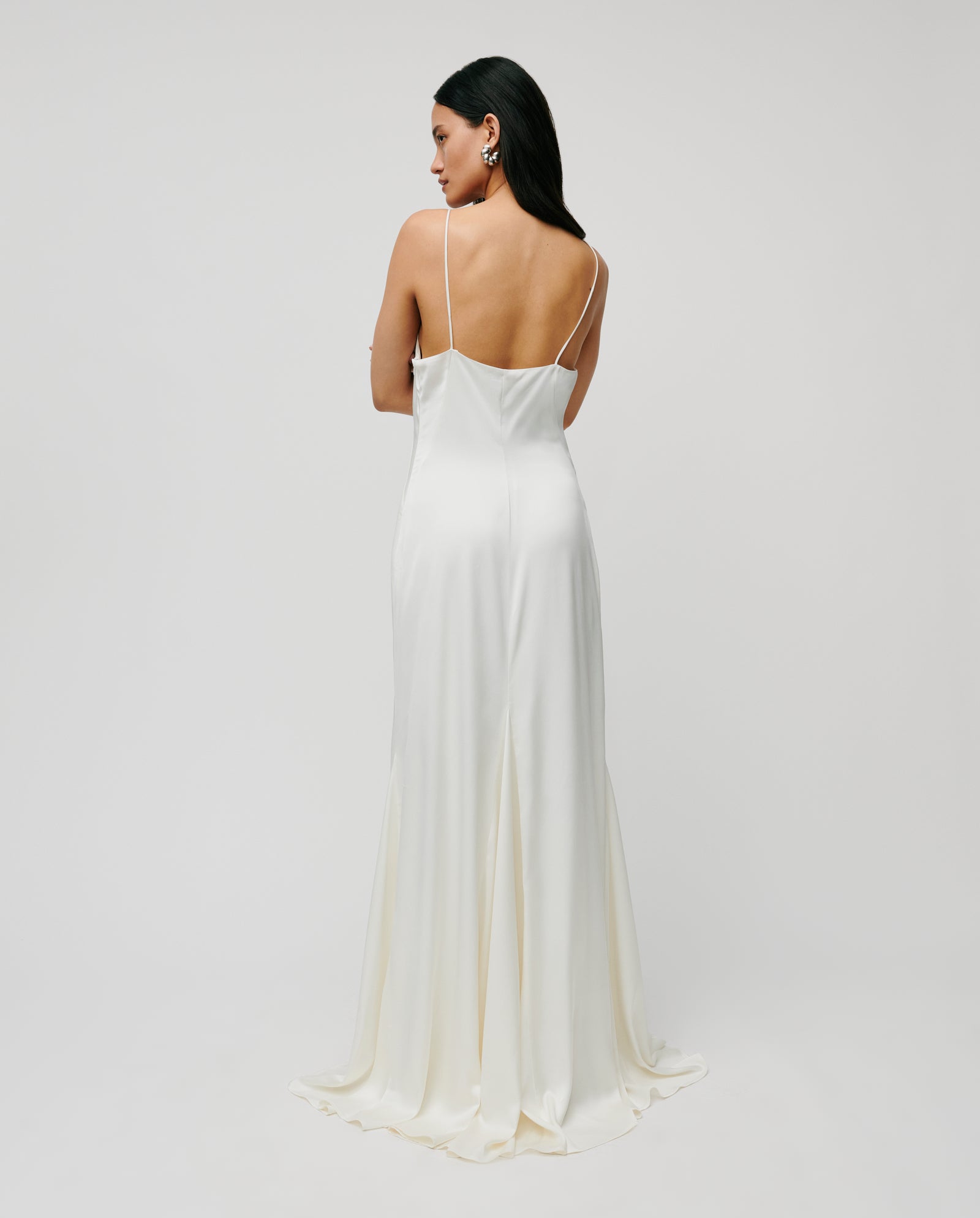 A woman stands with her back to the camera against a plain background, wearing the IVY OAK MADELIN IDA Bridaldress, a white satin NAIA acetate spaghetti strap gown.