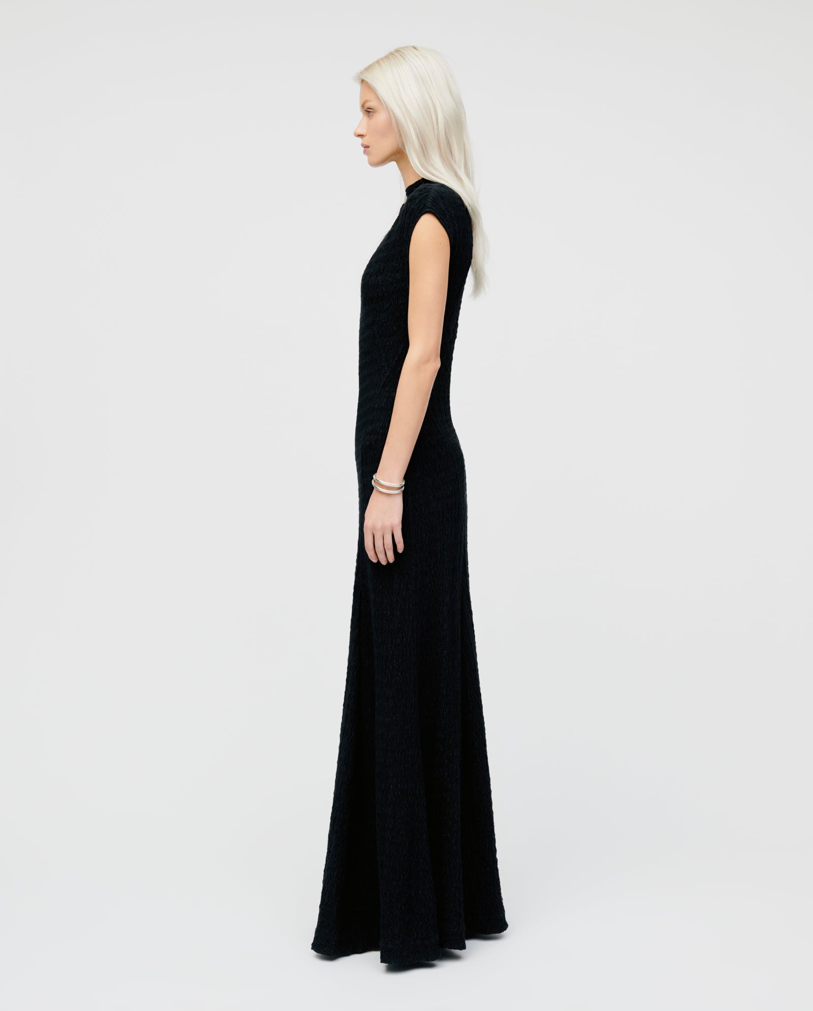 A woman stands in profile against a white background, wearing the IVY OAK MARCELLA Dress—an ankle-length, body-hugging black dress with short sleeves.