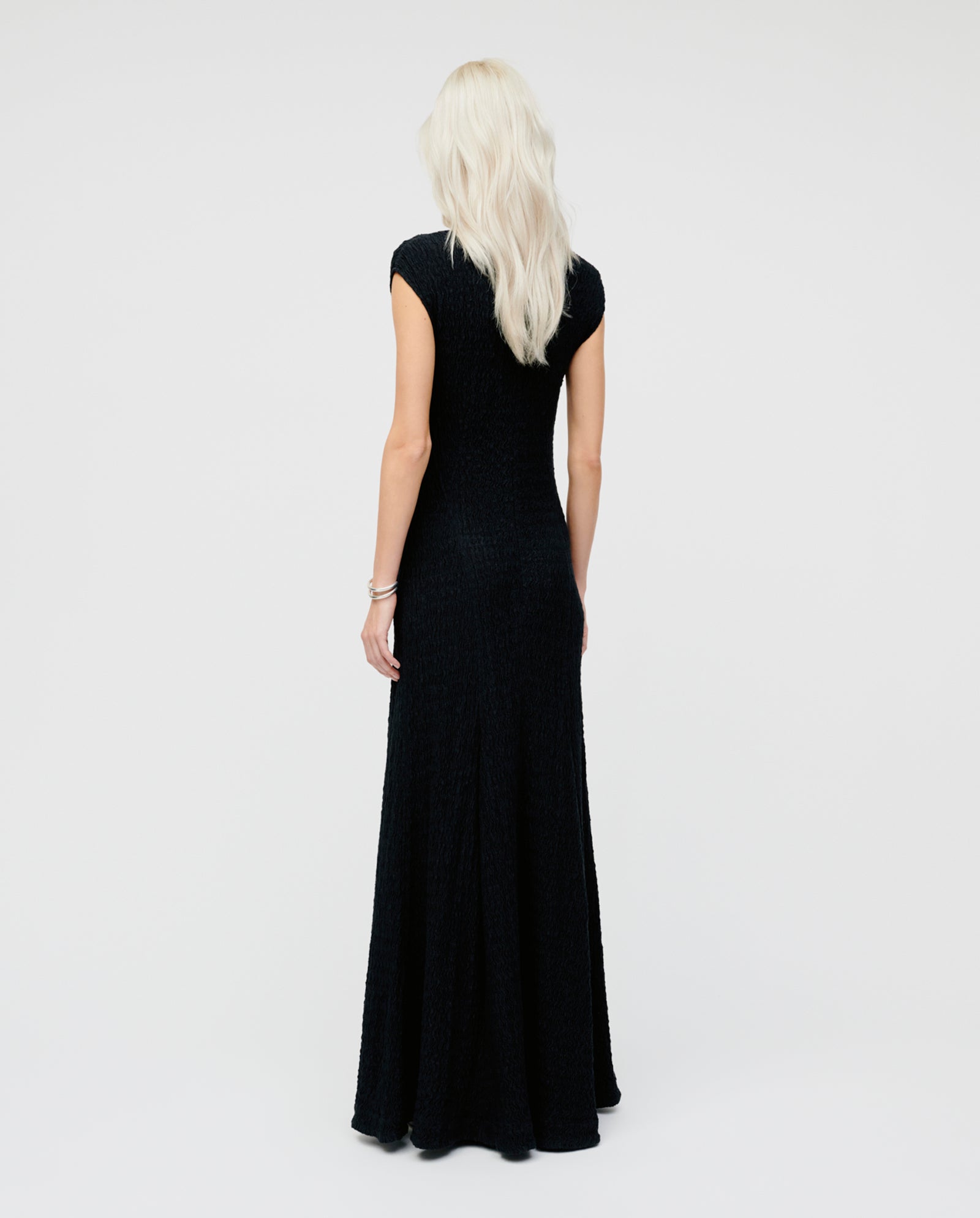 A woman with long blonde hair stands facing away, wearing the IVY OAK MARCELLA dress—a floor-length, body-hugging black gown with a flowing skirt.
