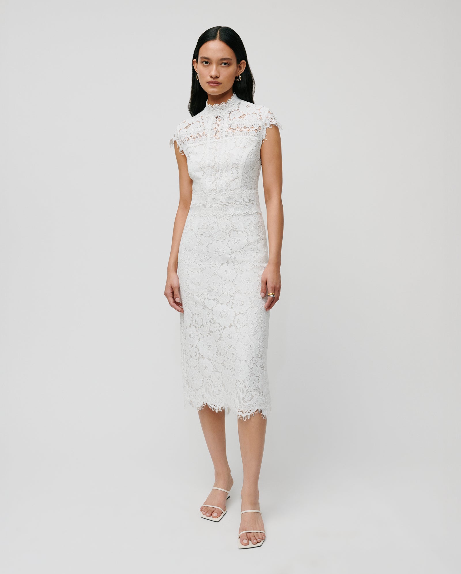 A woman models the IVY OAK NYNA ROSE Bridaldress—a fitted, white lace wedding dress with cap sleeves and a midi-length pencil skirt—against a plain background.