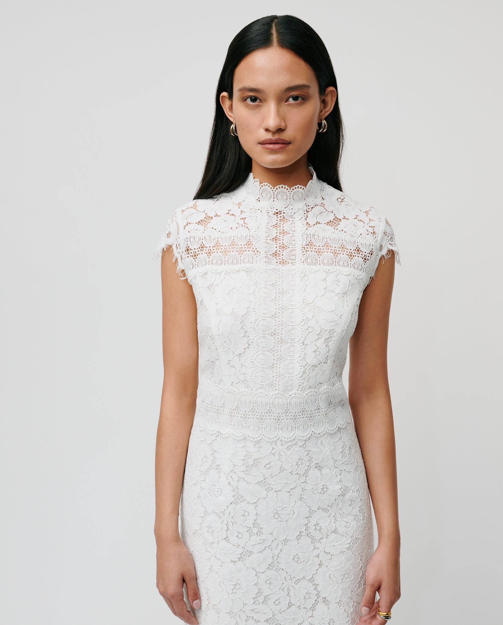 A woman wears the IVY OAK NYNA ROSE Bridal dress, featuring white lace, cap sleeves, and a midi length, standing against a plain light background.