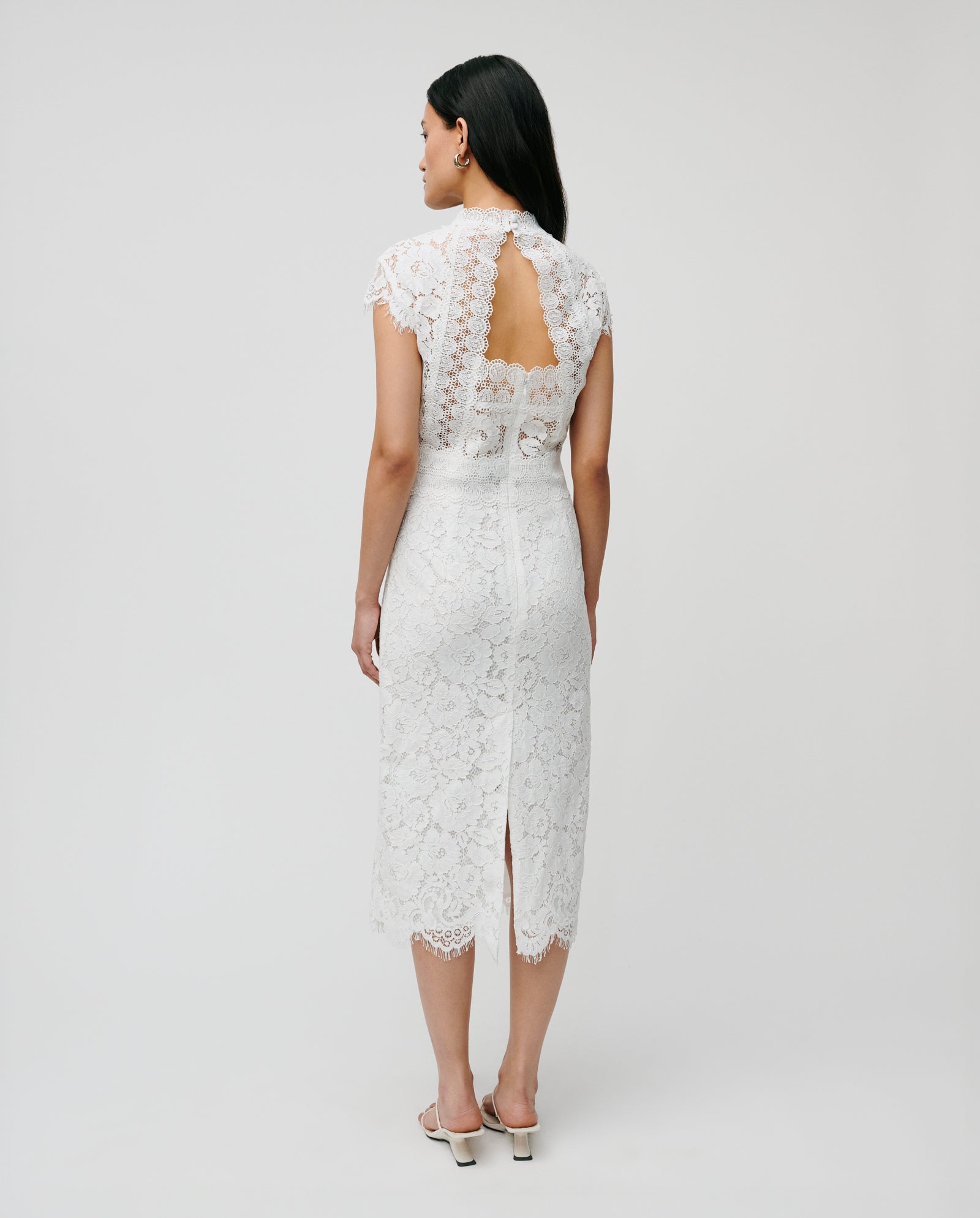 The NYNA ROSE Bridaldress by IVY OAK features a white lace design with cap sleeves, an open back, and an elegant midi length, worn by a woman facing away.