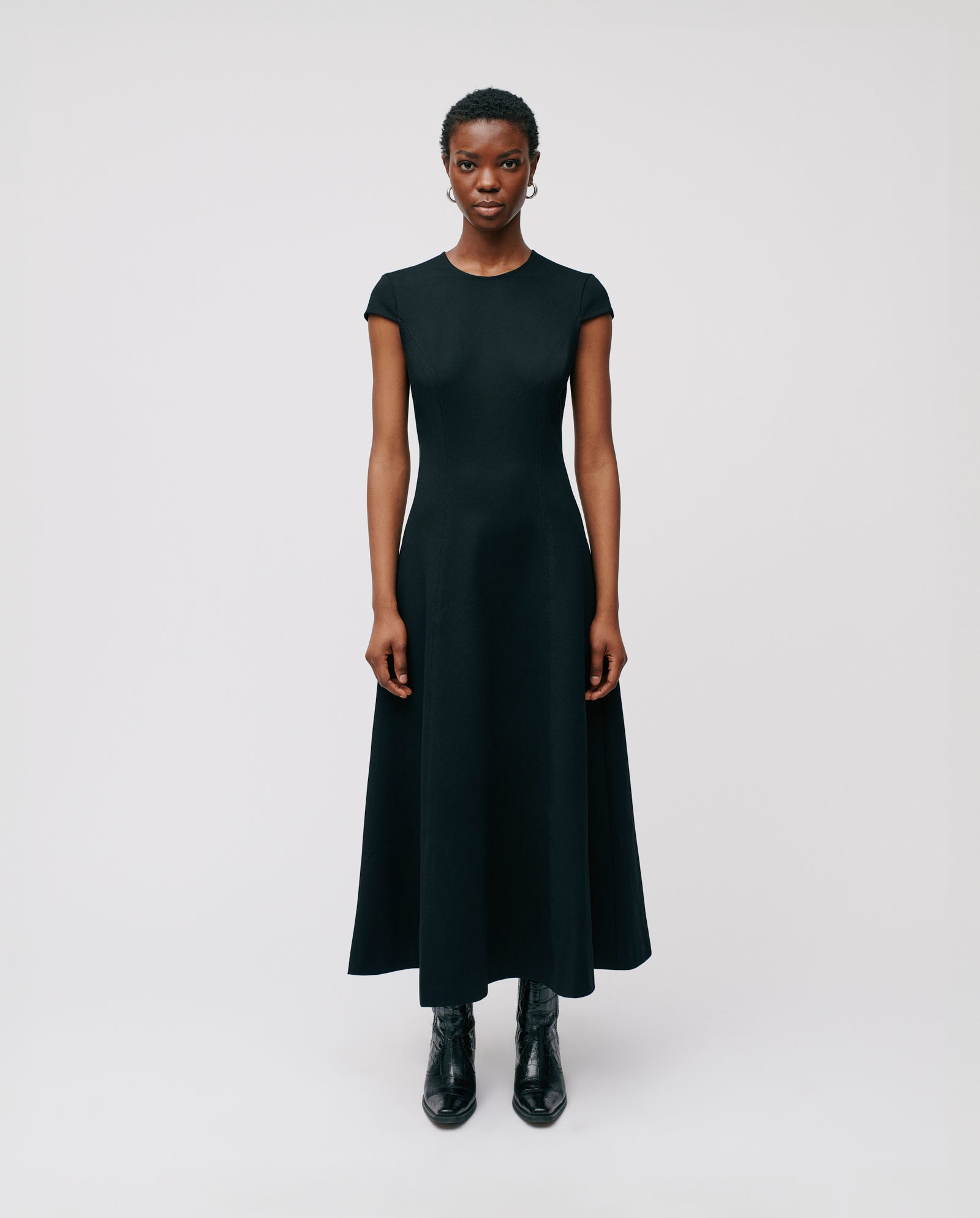 Woman in a black IVY OAK DAFNI Dress with short sleeves and ankle length, paired with black boots, stands against a white background—perfect for special occasions.