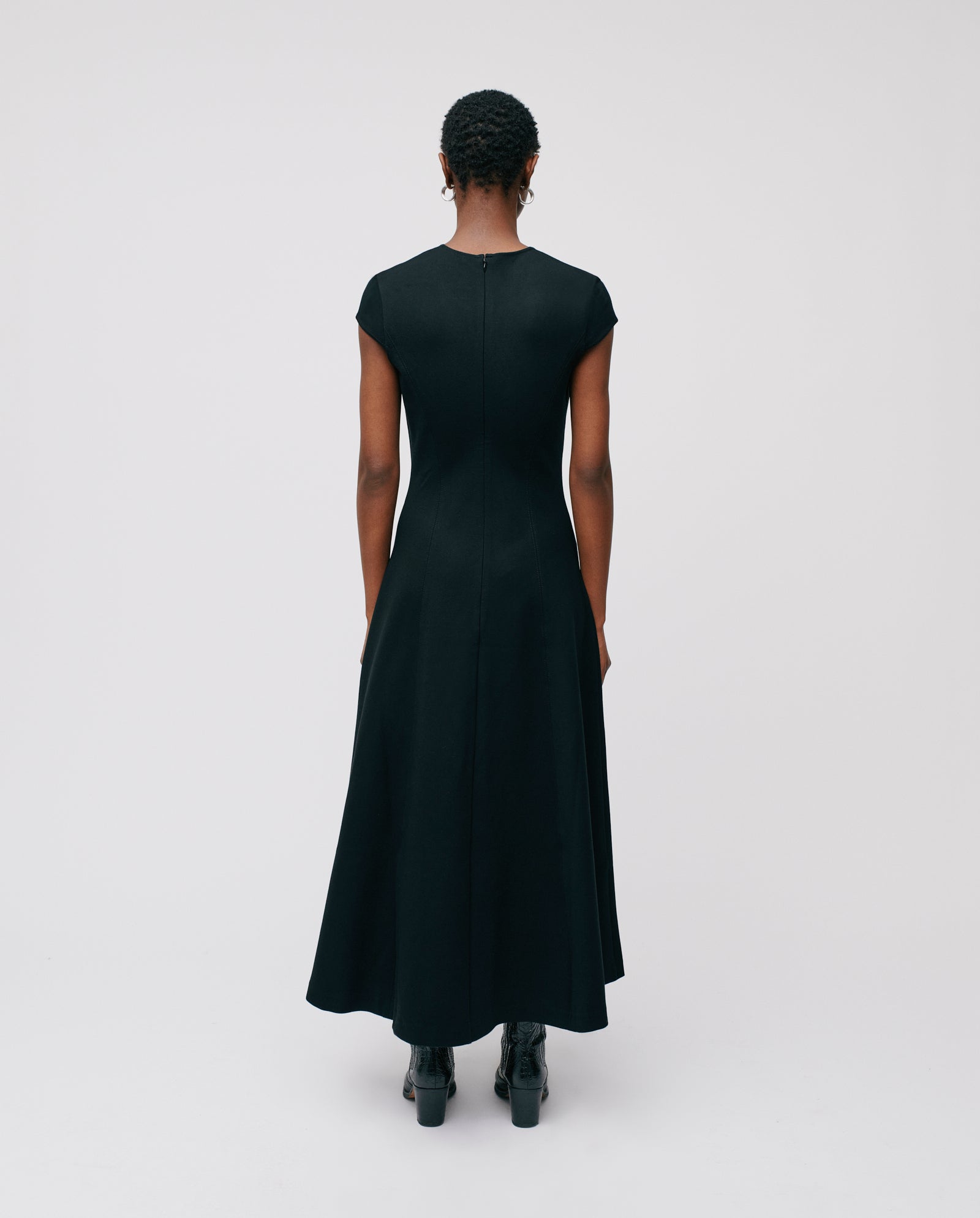 Person with short hair wears the IVY OAK DAFNI Dress, a black ankle-length outfit paired with boots, facing away against a plain background—a timeless style ideal for special occasions.