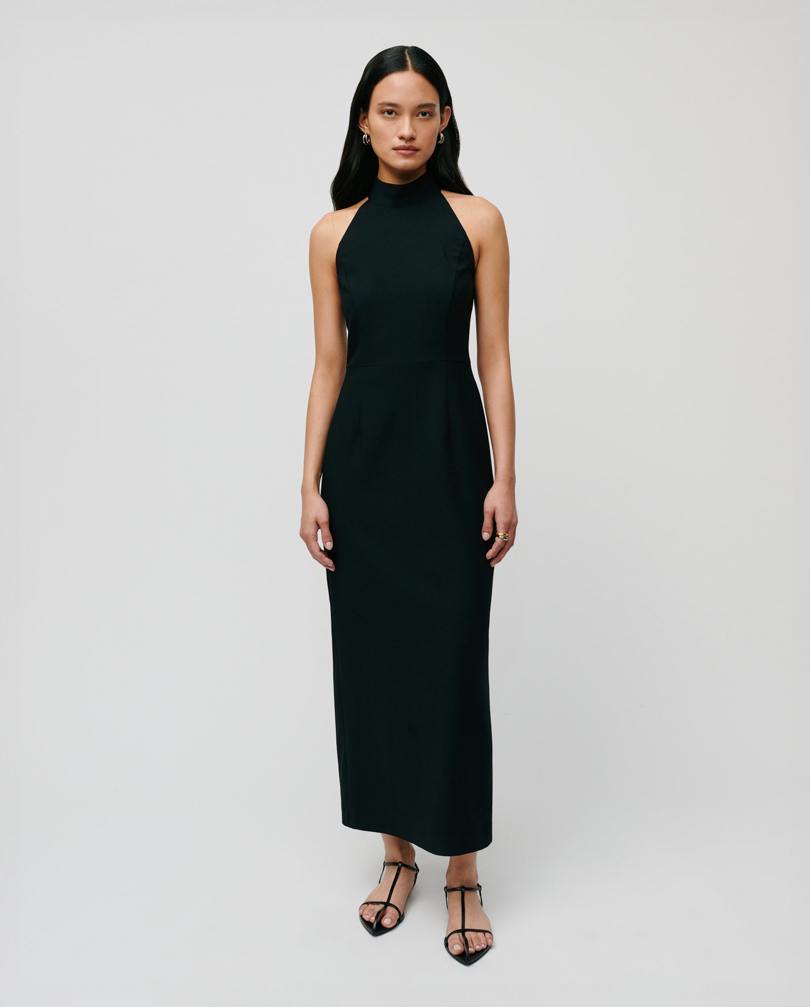 A woman wears the NOLAN Dress by IVY OAK—a sleeveless black neckholder dress with a body-hugging fit—paired with black strappy sandals, standing against a plain background.