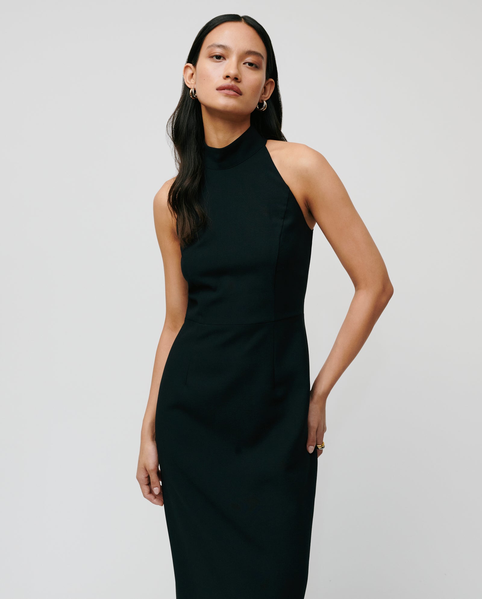 A woman models the NOLAN Dress by IVY OAK, a sleeveless, high-neck black dress with a body-hugging fit, standing against a plain light background.