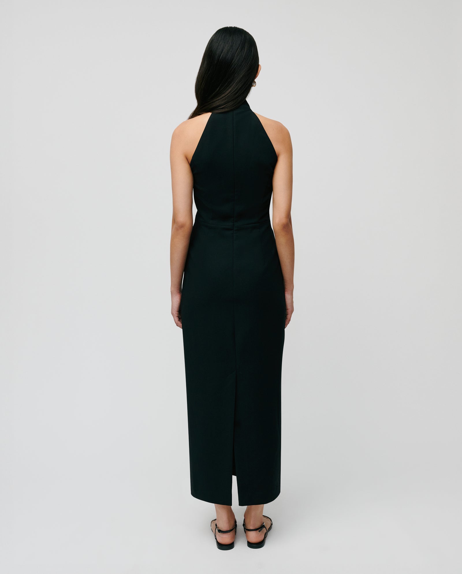 A woman stands with her back to the camera against a plain background, wearing the IVY OAK NOLAN Dress—a sleeveless, body-hugging black neckholder style—paired with sandals.