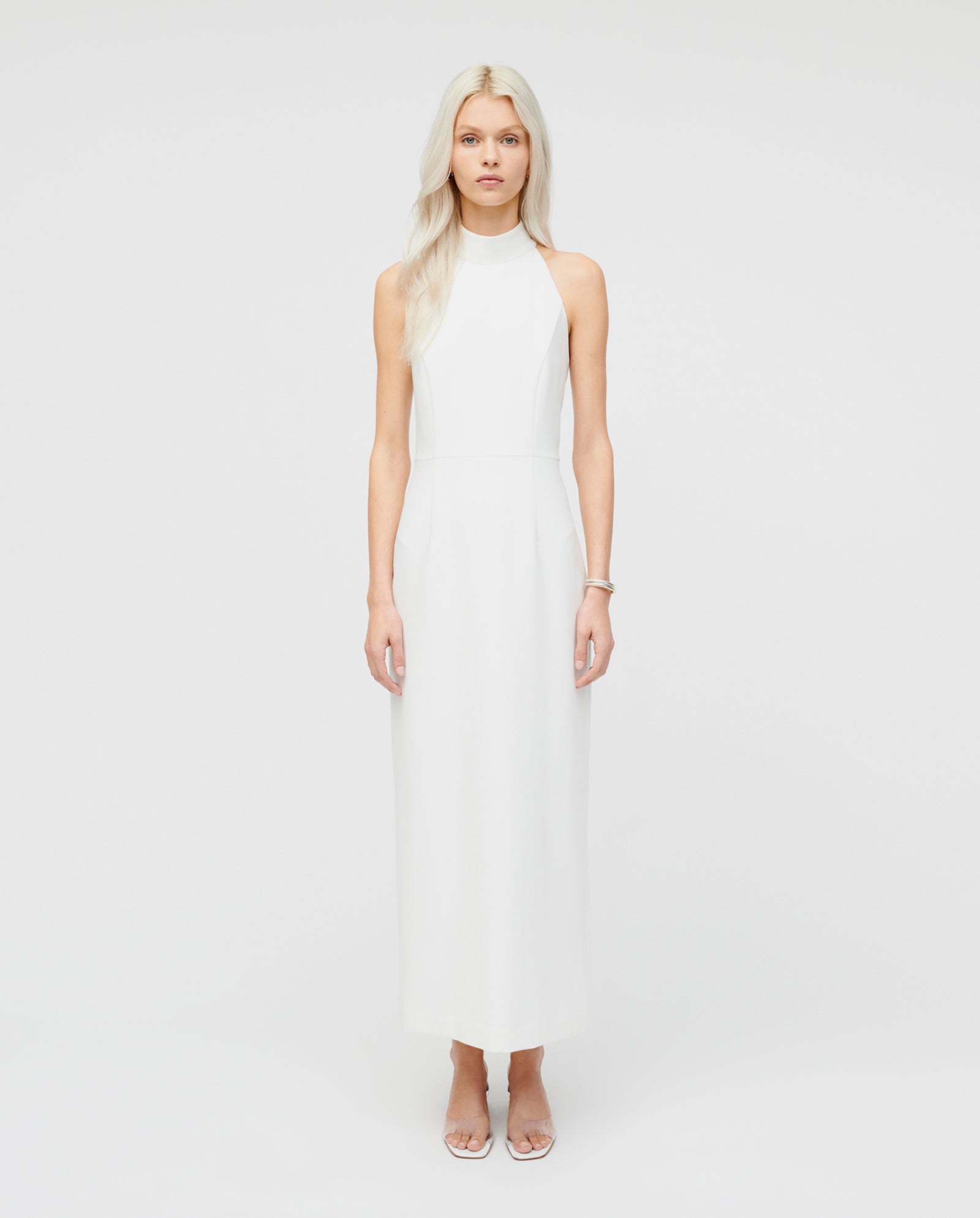 A woman with long blonde hair wears an ankle-length IVY OAK NOLAN Eveningdress and clear heels, standing in a studio.