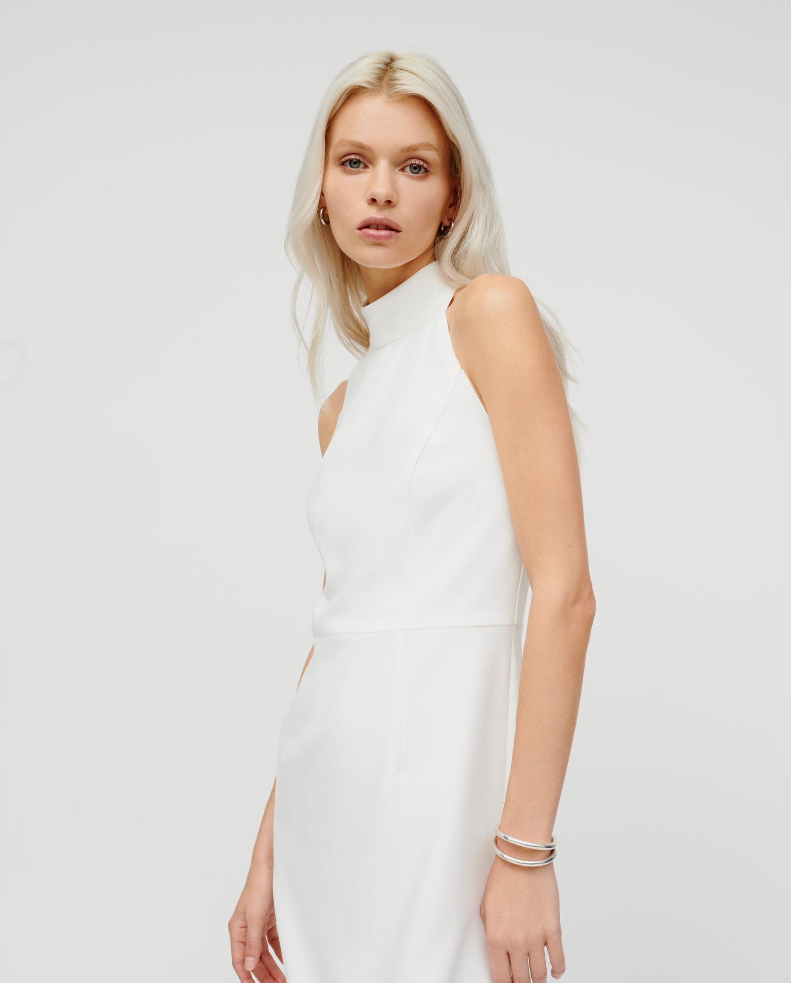Woman with long blond hair poses against a plain light background, wearing the IVY OAK NOLAN Dress—a sleeveless, high-neck white neckholder style.