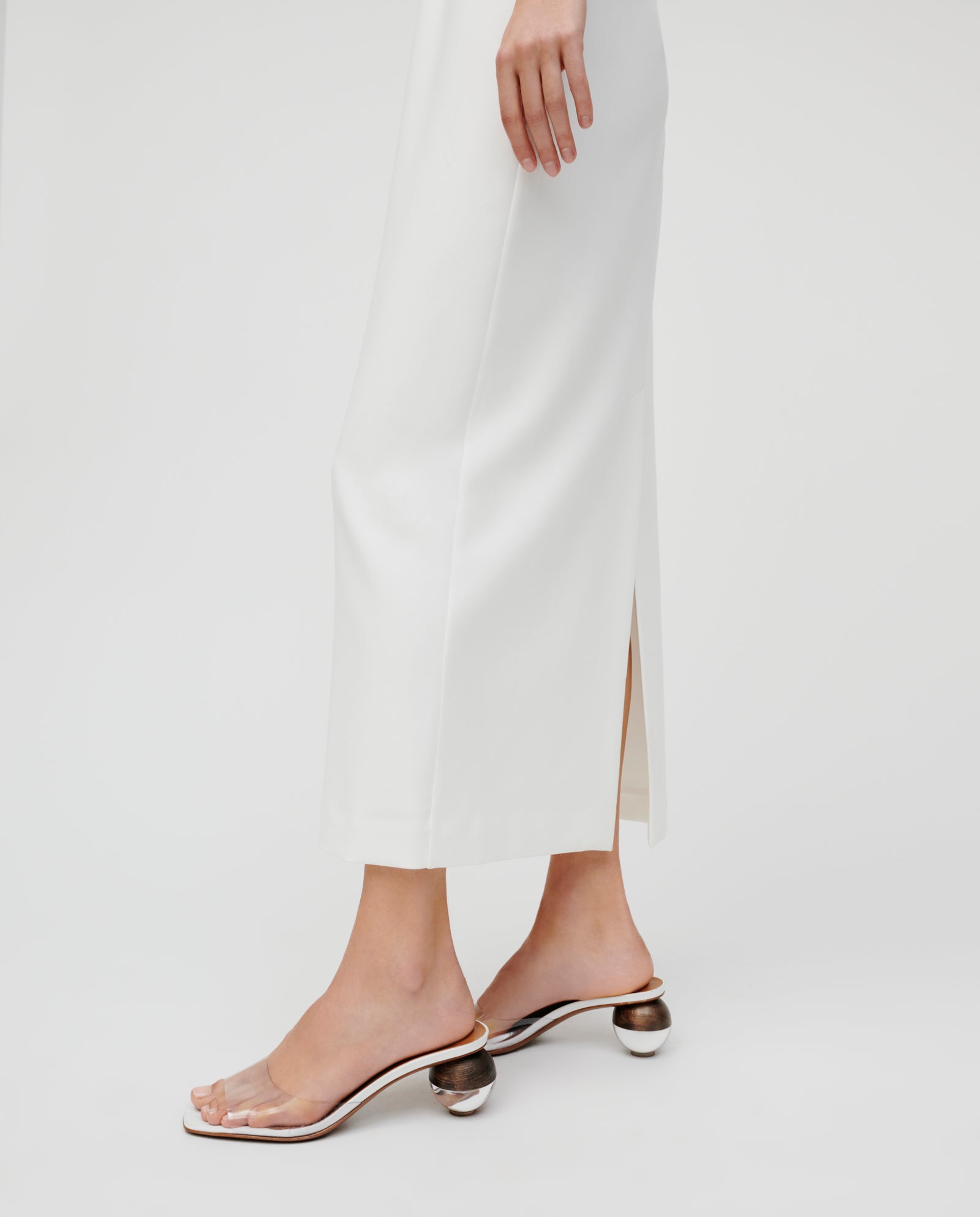 A person in the IVY OAK NOLAN Eveningdress, an ankle-length, body-hugging white skirt, styled with clear heeled sandals, stands against a plain white background.