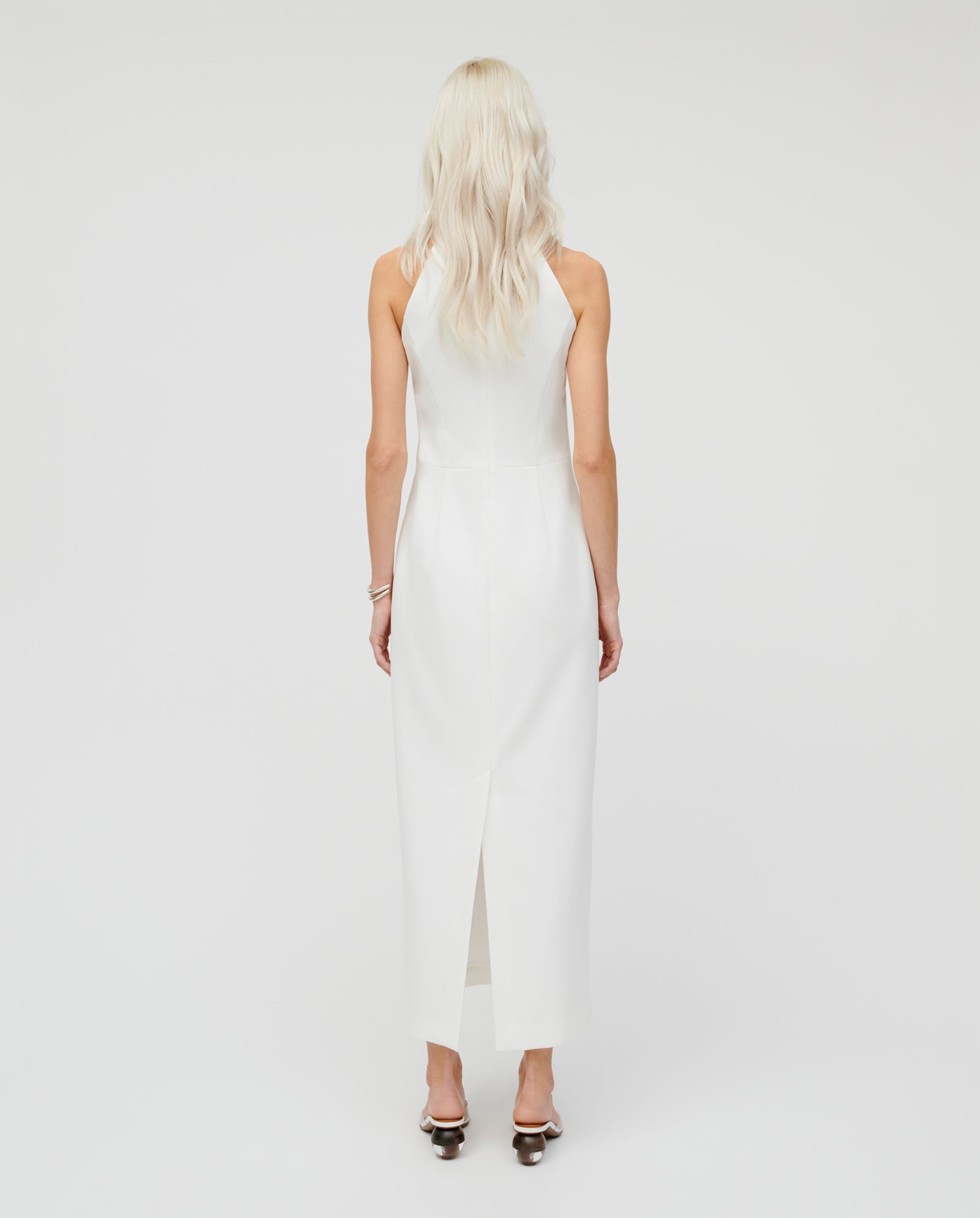 A woman with long blonde hair models the IVY OAK NOLAN Eveningdress, showcasing its fitted, ankle-length silhouette from the back against a white background.