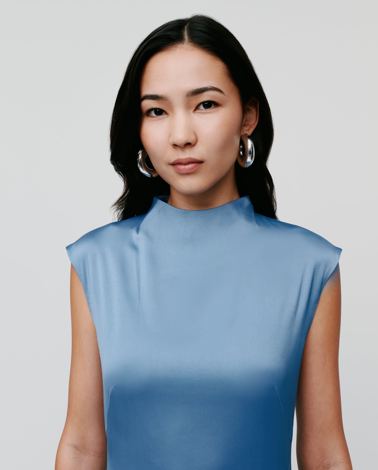 Woman with long dark hair wearing the IVY OAK MARCELLA LIZ Dress, a sleeveless blue design with a stand-up collar and accessorized with large hoop earrings, facing forward.