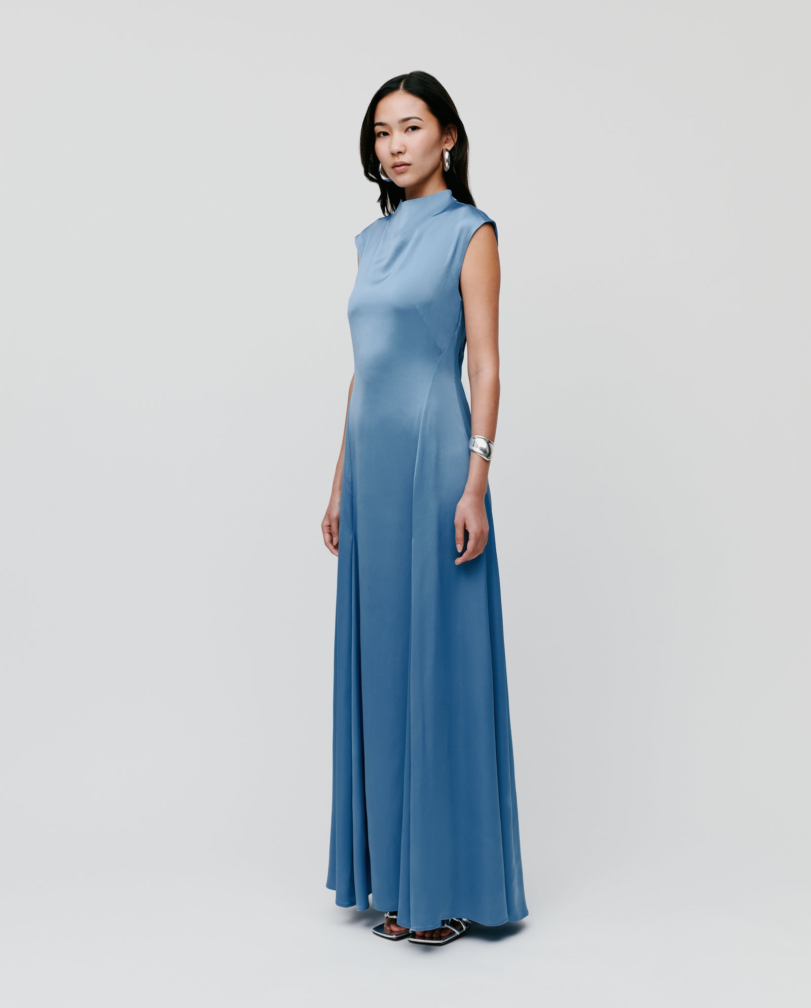 A woman wears the IVY OAK MARCELLA LIZ sleeveless, floor-length blue maxi dress with a stand-up collar, standing against a plain white background.
