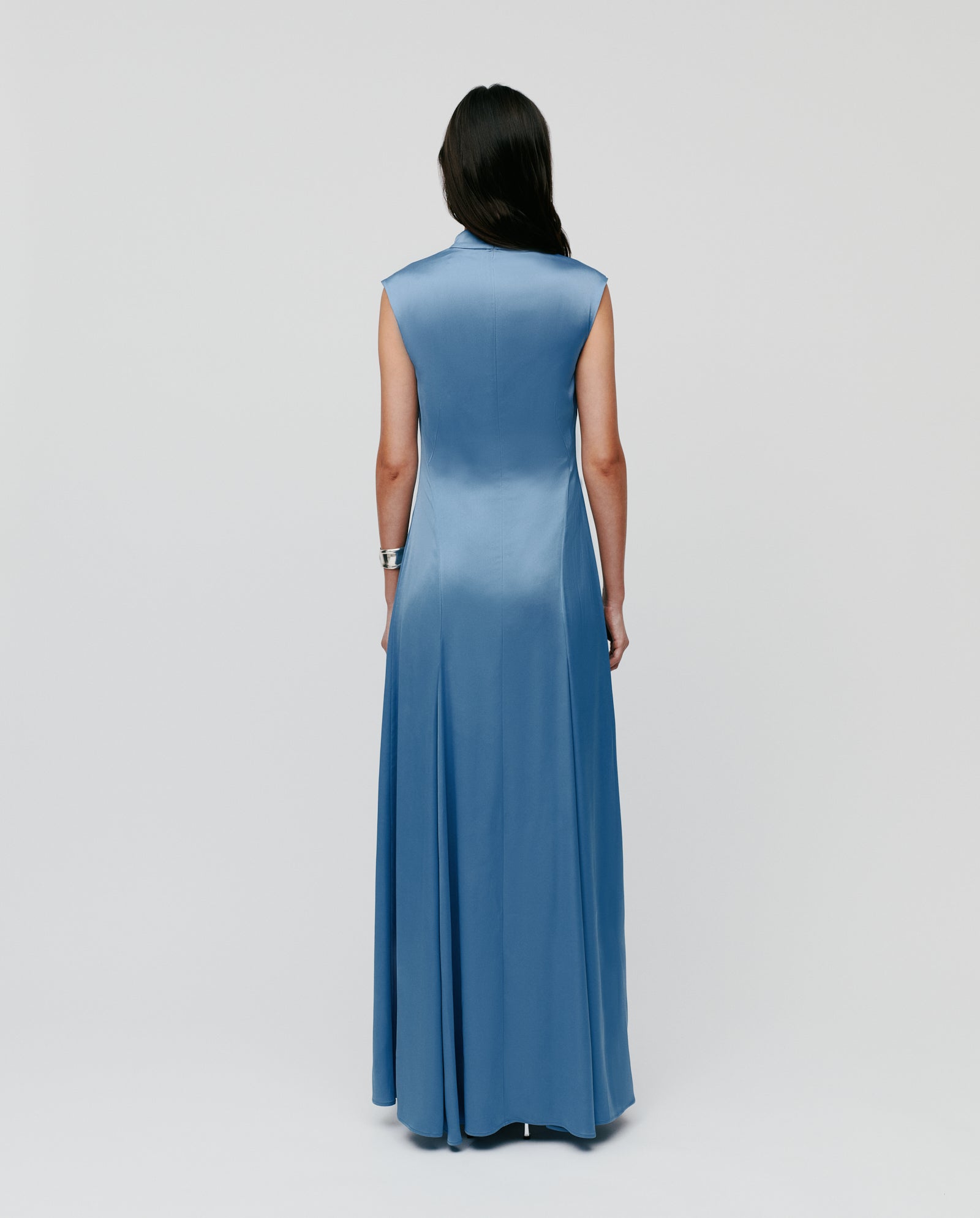 A woman wearing the IVY OAK MARCELLA LIZ Dress—a sleeveless blue maxi with a stand-up collar and back zipper—stands facing away against a plain light background.