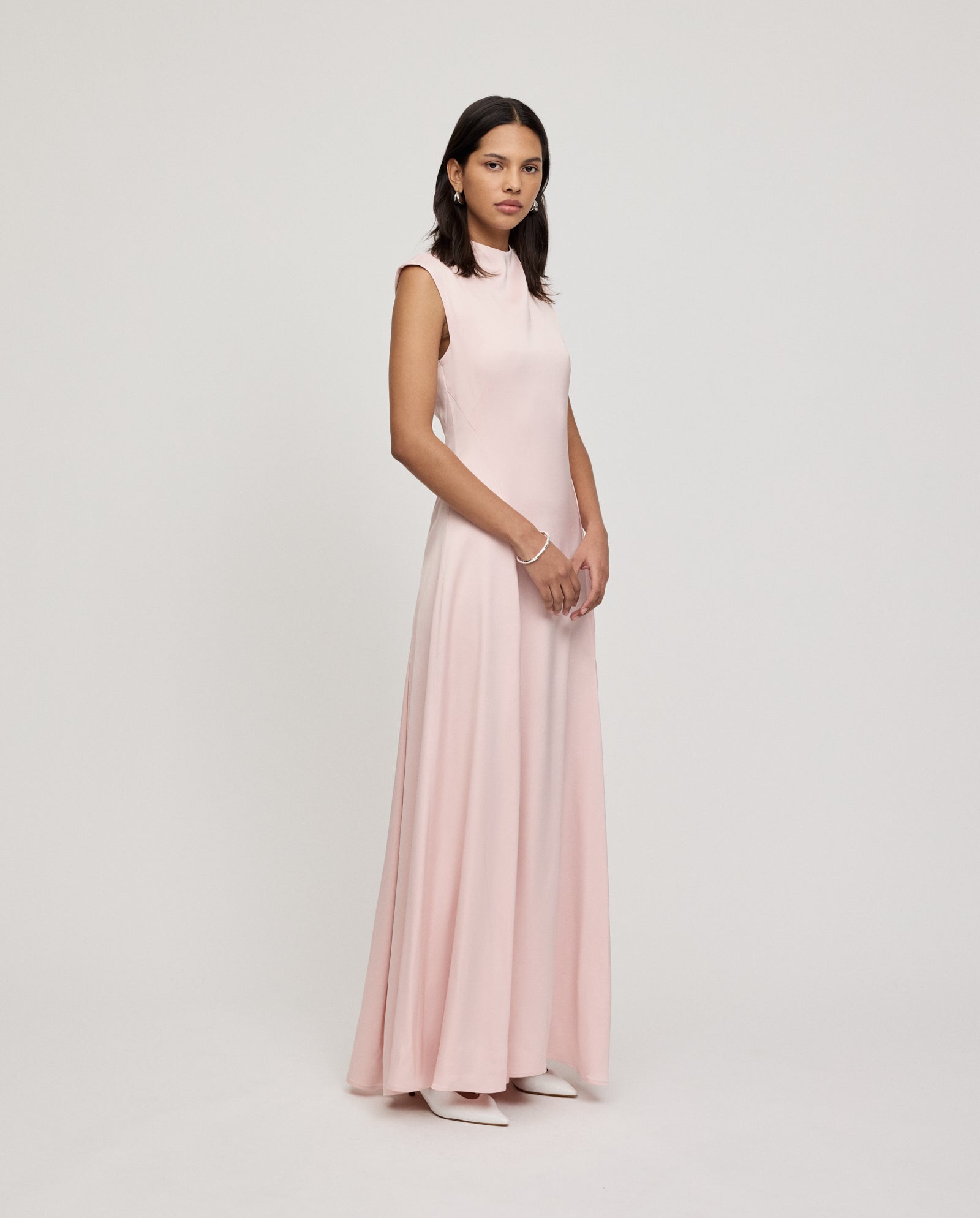 A woman wears the IVY OAK MARCELLA LIZ dress, a sleeveless, floor-length pale pink maxi with godets, standing against a plain white background.