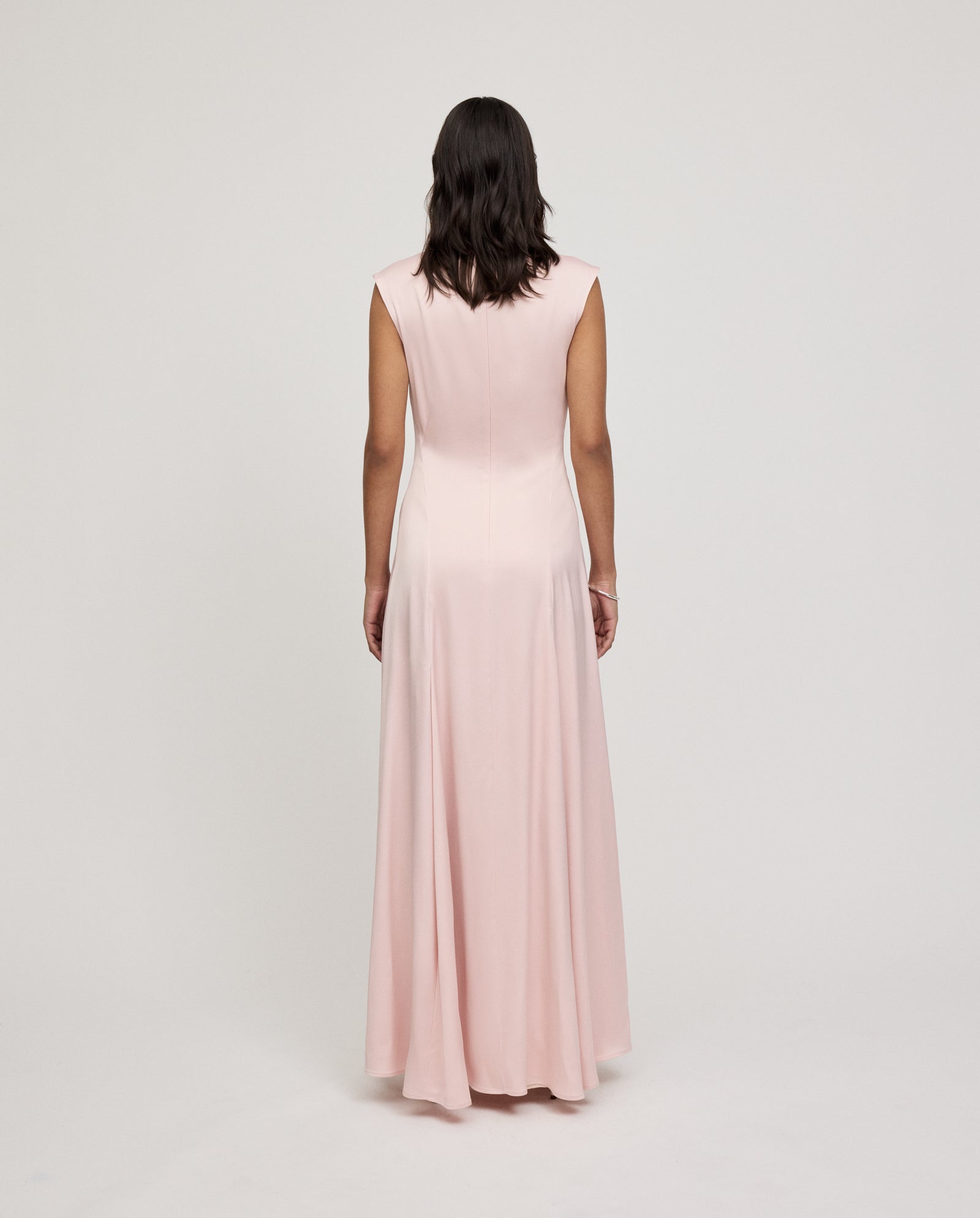 A person wearing the IVY OAK MARCELLA LIZ Dress—a sleeveless, floor-length light pink maxi with subtle godets—stands facing away against a plain background.