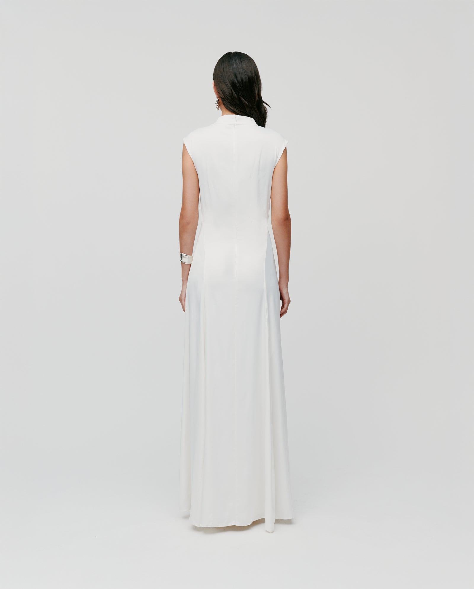 Woman in a sleeveless MARCELLA LIZ Dress by IVY OAK stands with her back to the camera, a plain background emphasizing the elegant godets in the skirt and its chic stand-up collar.