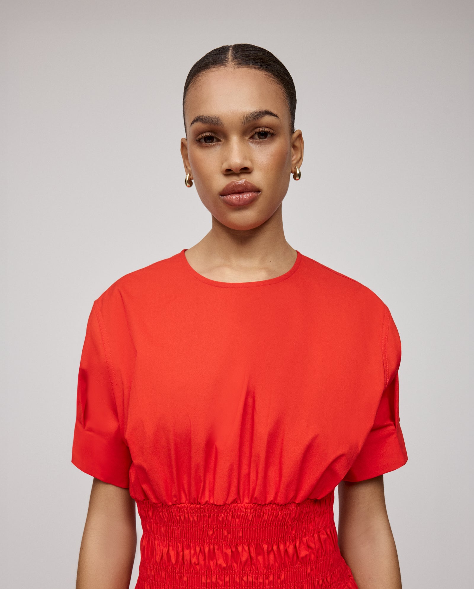 A woman wearing a red, V-neck MARYTHA Dress by IVY OAK with short sleeves, gold hoop earrings, and her hair pulled back stands against a plain background.