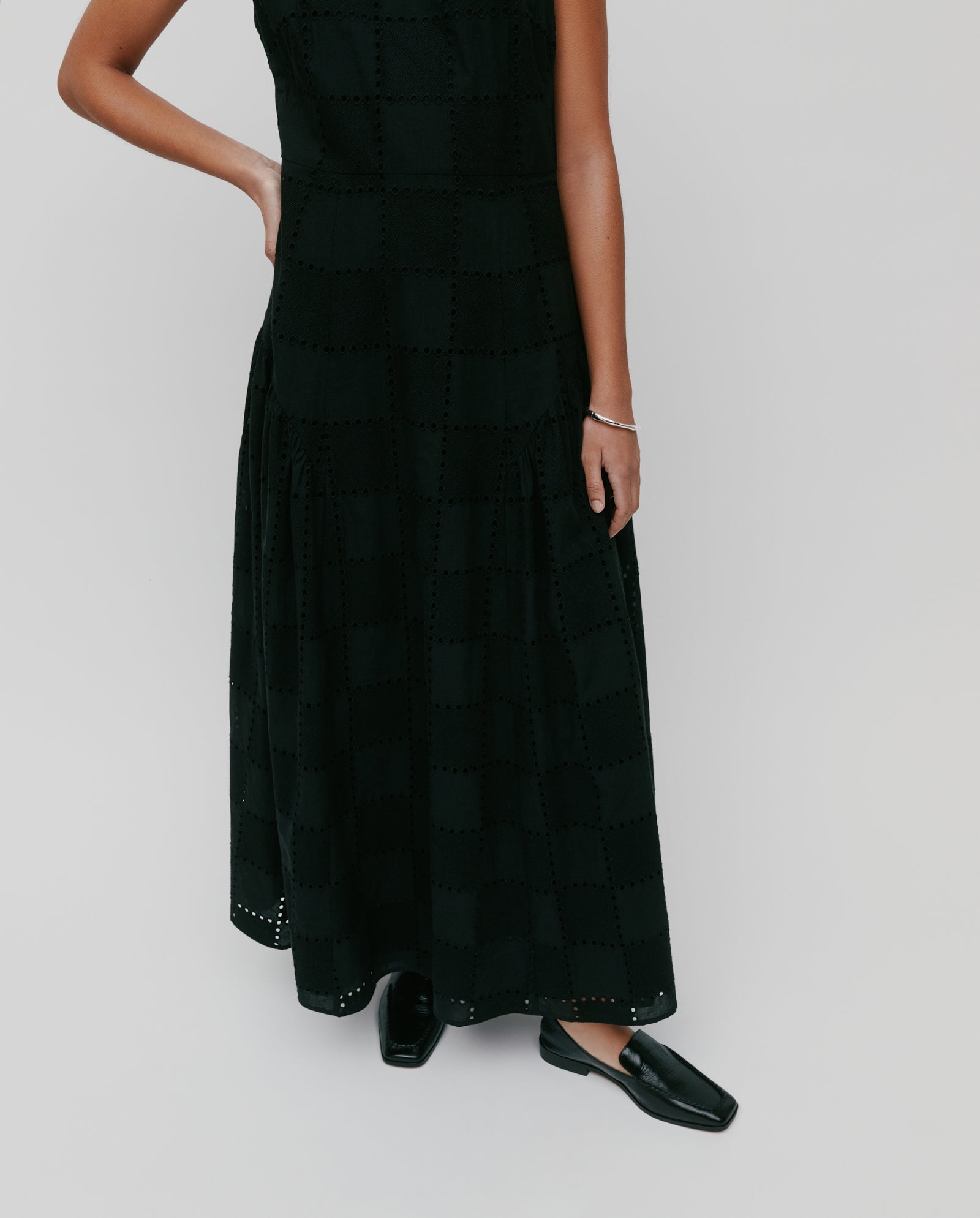 Woman in the IVY OAK MARCA ROSE sleeveless black maxi dress with a gathered skirt and black shoes, standing with one hand on her hip.