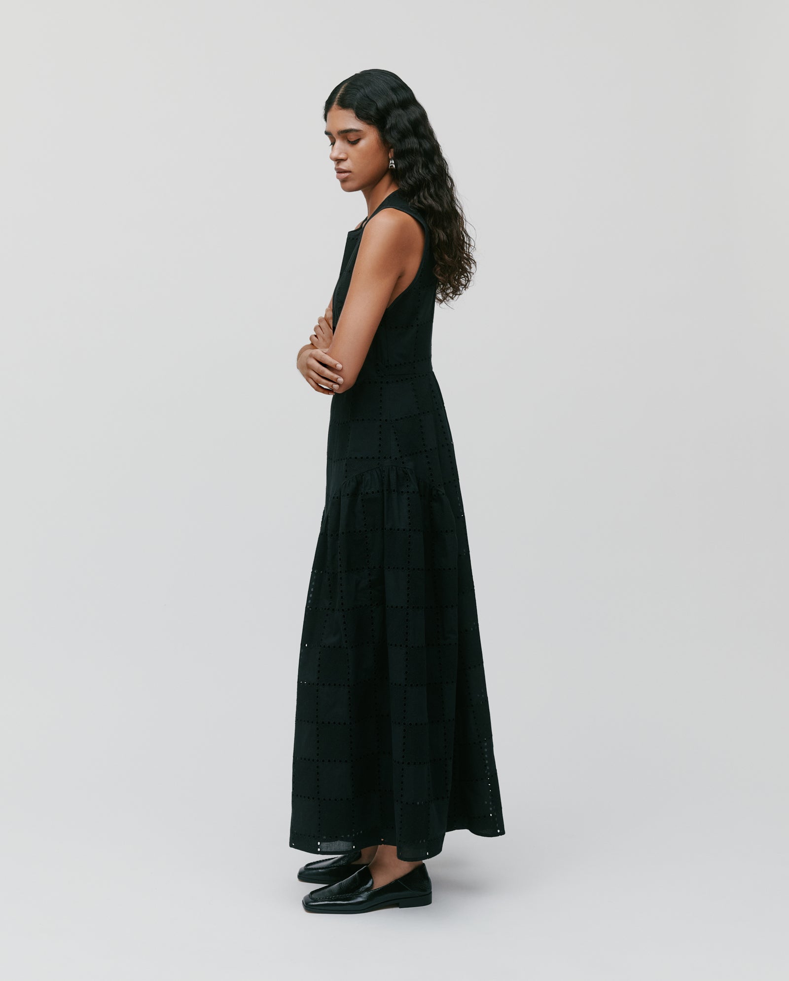 A woman stands sideways against a plain light background wearing the IVY OAK MARCA ROSE long black romantic dress with a gathered skirt and black shoes.