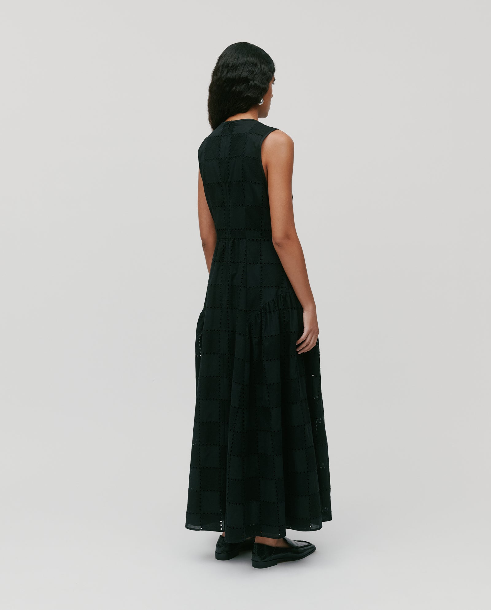 Woman in the MARCA ROSE dress by IVY OAK—a sleeveless, long black romantic dress featuring a subtle check pattern and softly gathered skirt—stands facing away.
