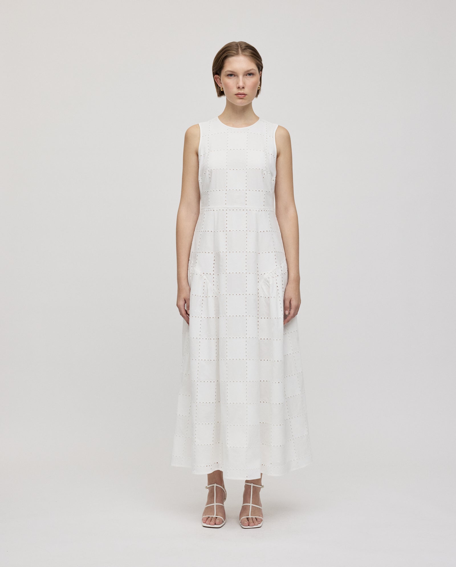 A woman stands still in the ankle-length, sleeveless MARCA ROSE Dress by IVY OAK, featuring romantic square-patterned broderie anglaise and a gathered skirt, paired with white strappy heels.