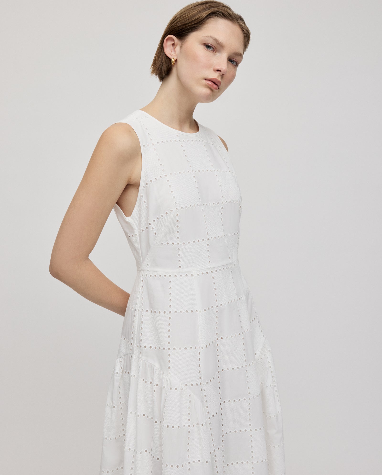 A woman poses against a plain background wearing the IVY OAK MARCA ROSE Dress, a sleeveless white broderie anglaise piece featuring geometric cutout patterns and gathered skirt inserts.