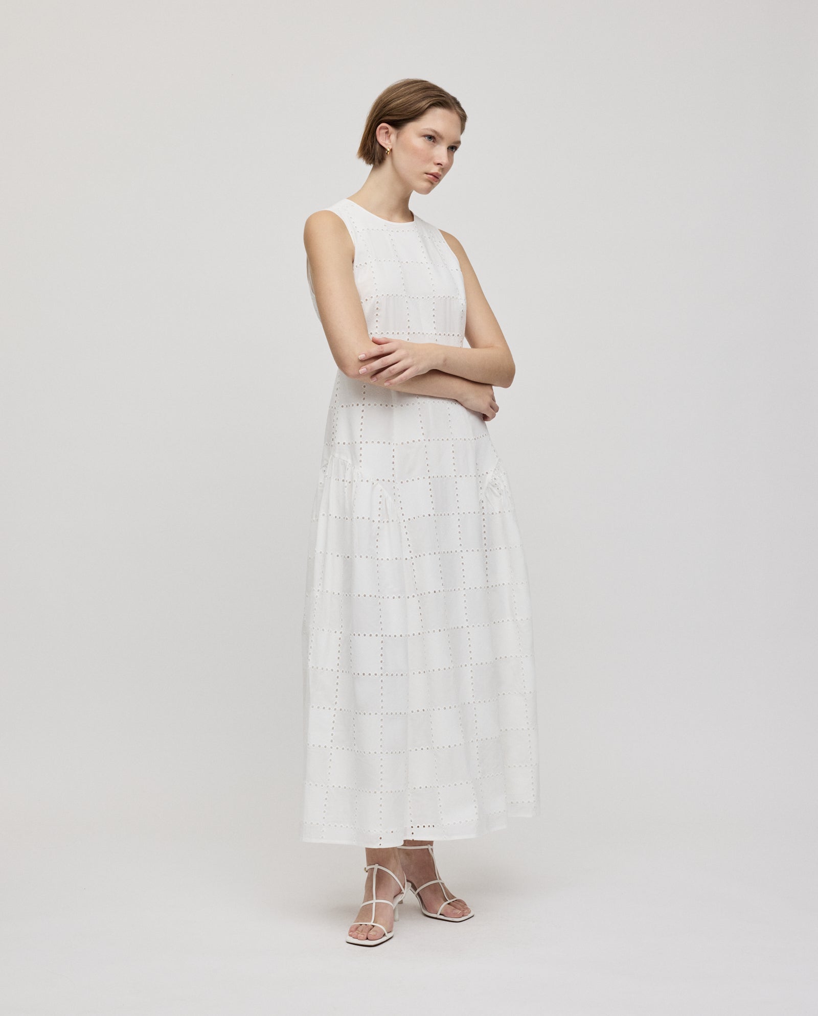 Woman in the IVY OAK MARCA ROSE sleeveless long white broderie anglaise dress with gathered skirt inserts and white sandals, standing with arms crossed.