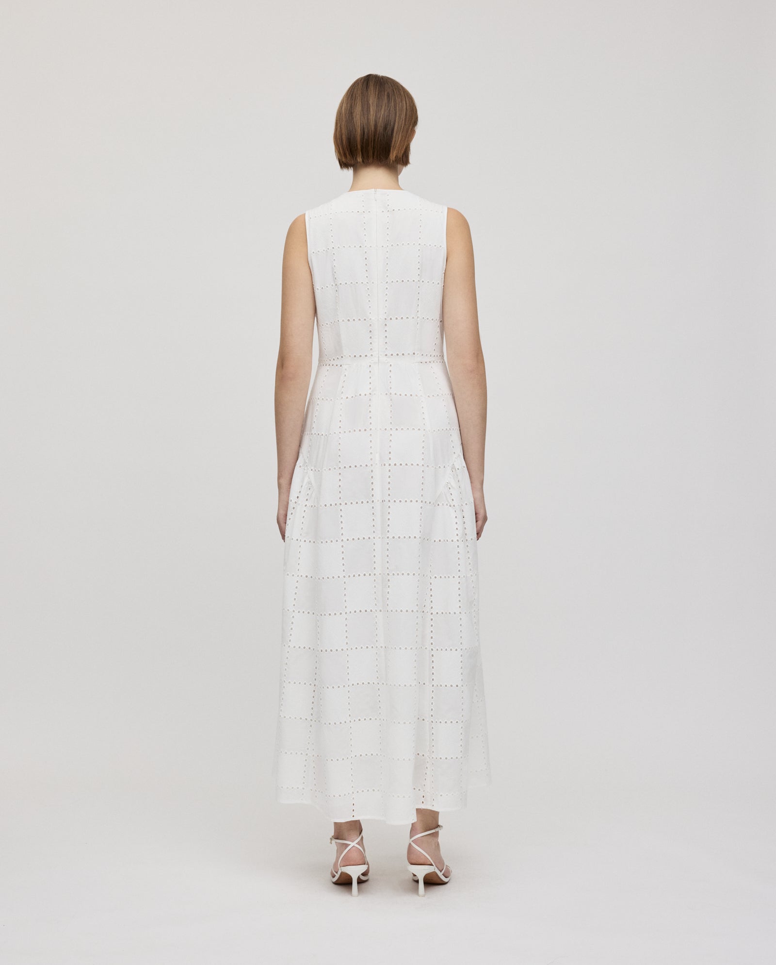 A woman stands with her back to the camera, wearing the IVY OAK MARCA ROSE sleeveless romantic dress featuring a gathered skirt and elegant white heels.