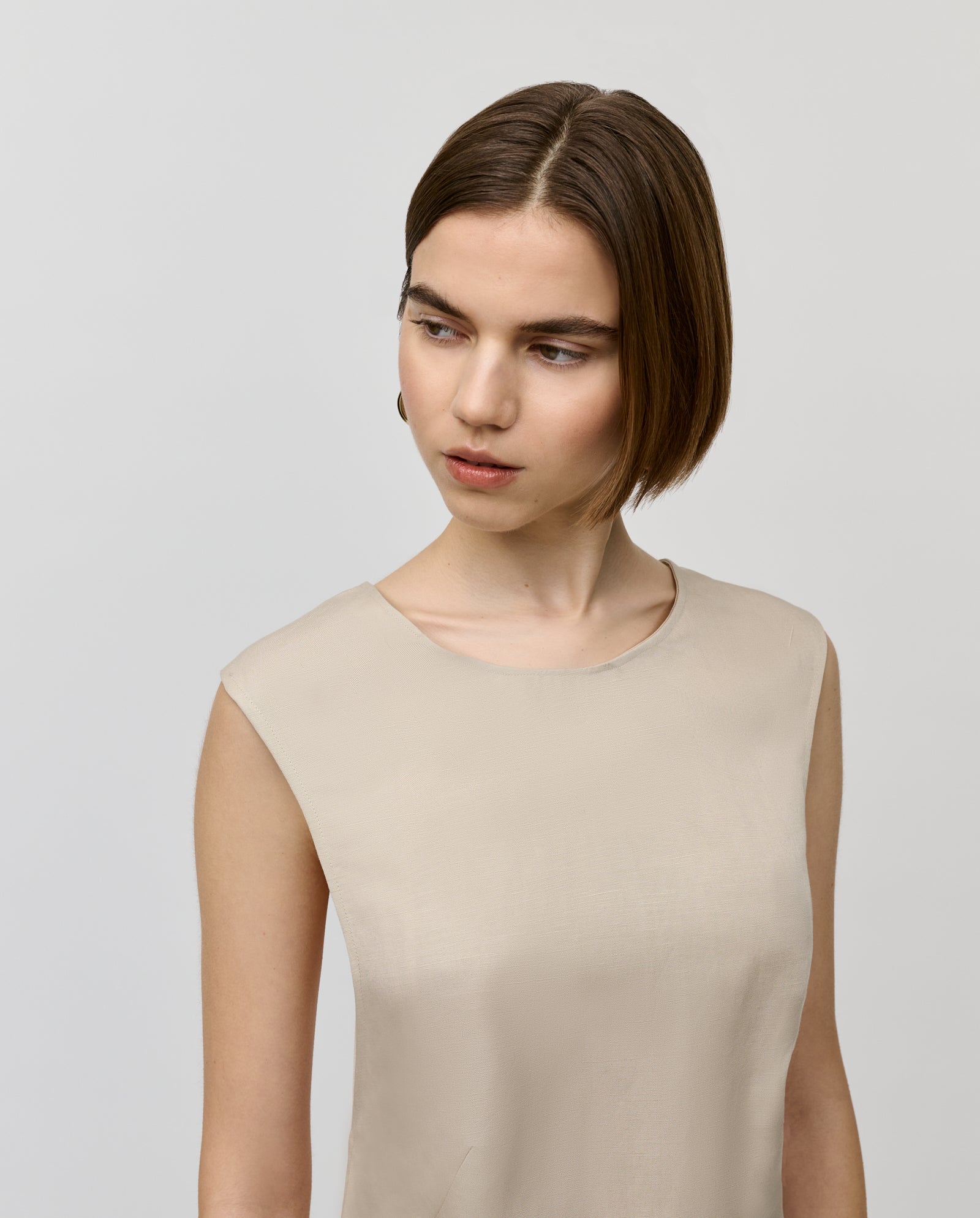 A woman in a sleeveless beige IVY OAK NADEAN Dress gazes thoughtfully to the side against a plain light background.