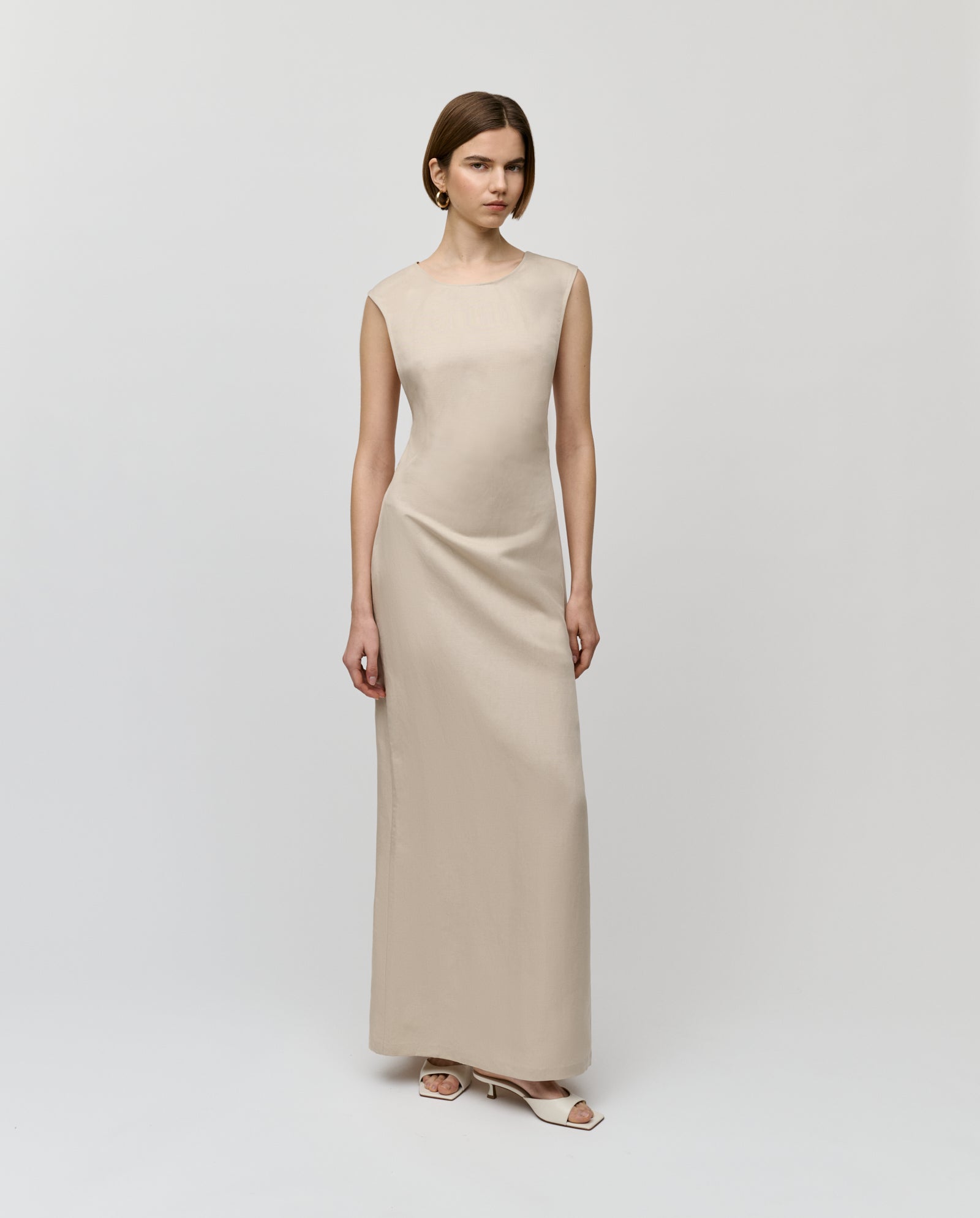 A woman wears the IVY OAK NADEAN Dress, a sleeveless beige linen-blend piece, paired with white heels and set against a plain light background.