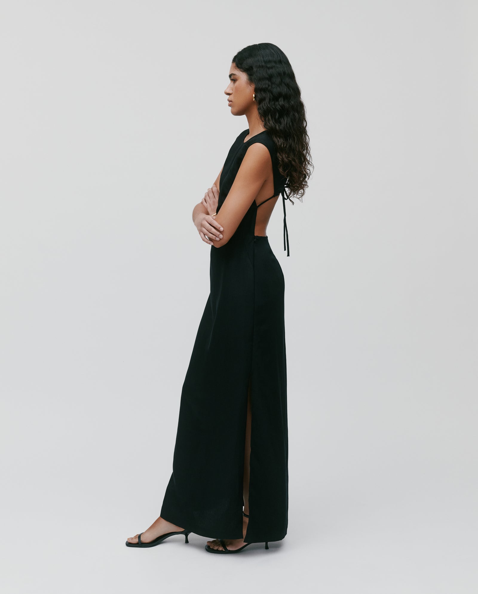 Woman in the NADEAN long, sleeveless linen blend black dress by IVY OAK with an open back, standing sideways with arms crossed.