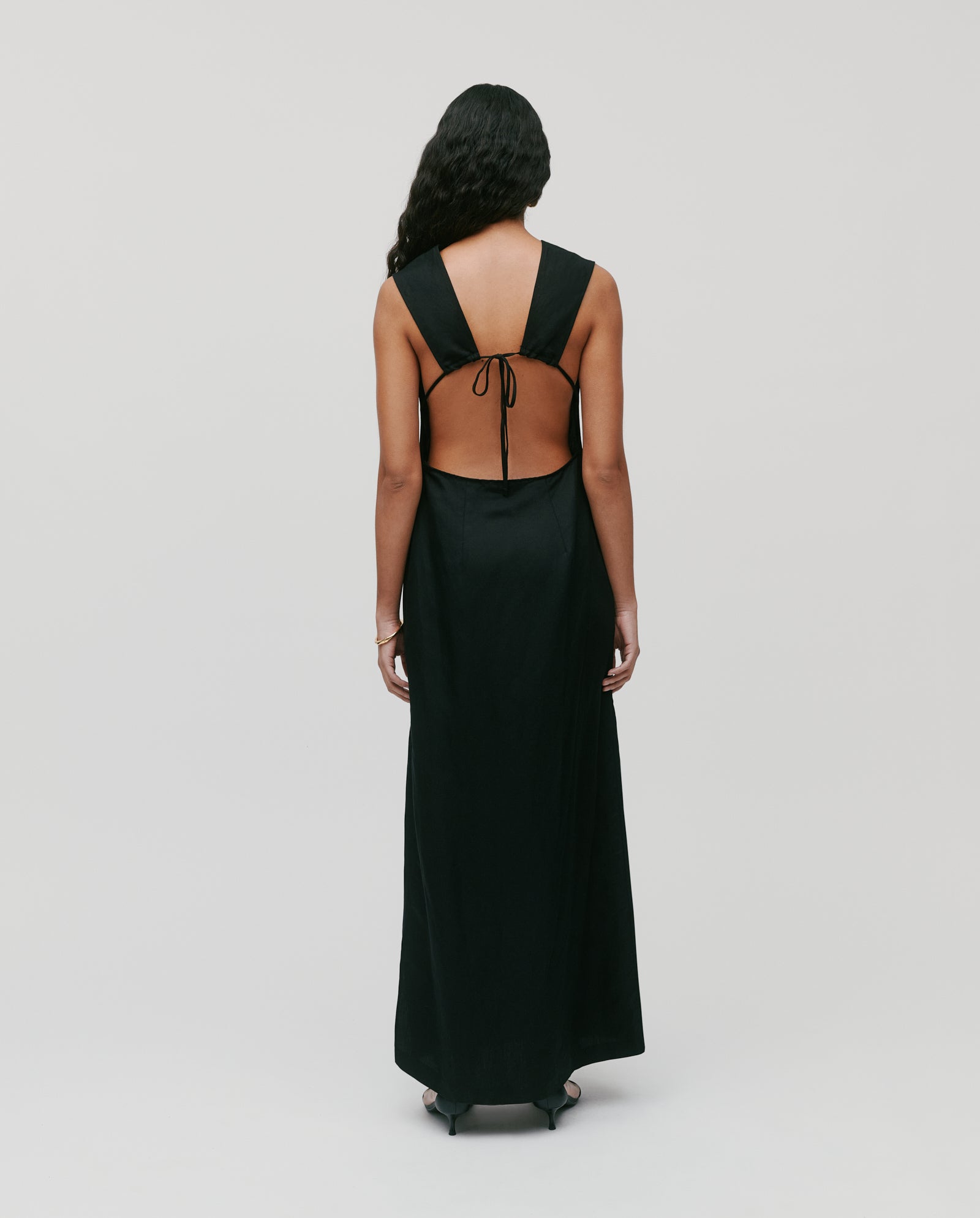 A woman in the NADEAN Dress by IVY OAK, an elegant linen blend piece with an open back and tie detail, stands facing away against a plain light background.