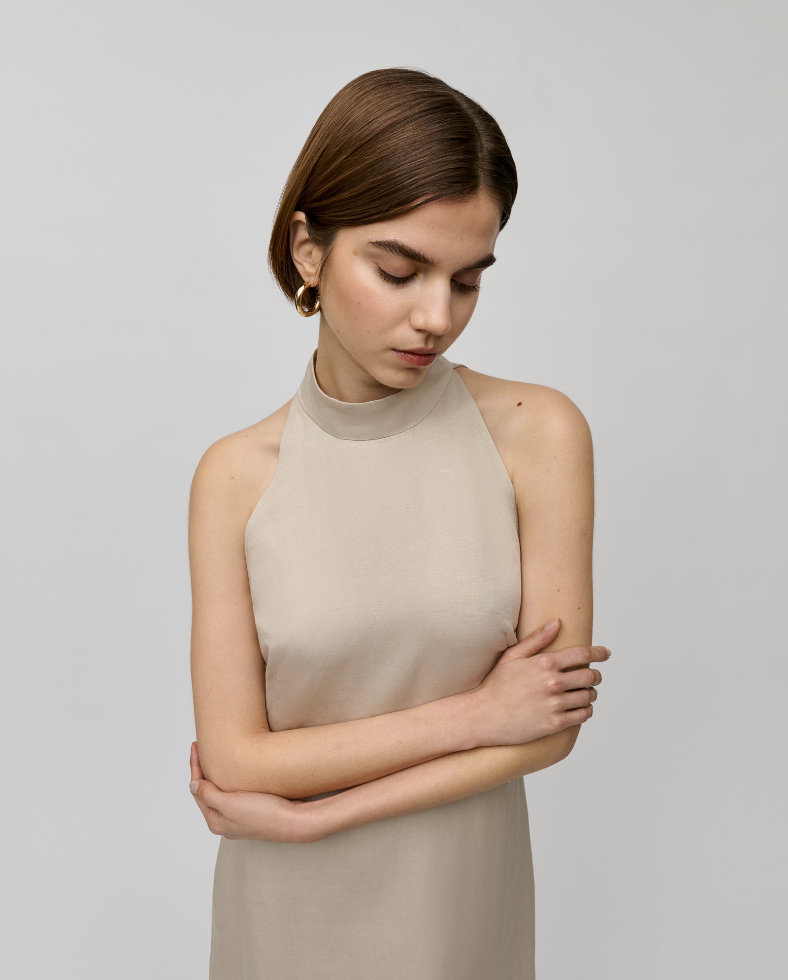 A woman with short brown hair stands with arms crossed, looking down, wearing the elegant NELA Dress by IVY OAK, crafted from a linen blend fabric.