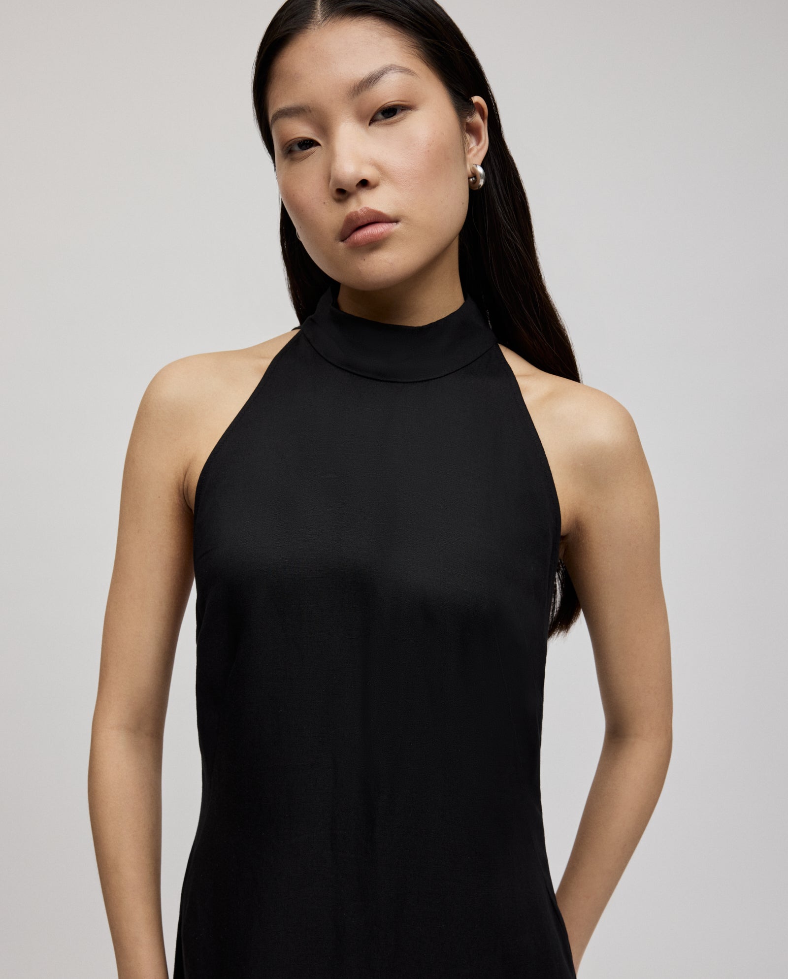 A woman with long dark hair wears the sleeveless black NELA Dress by IVY OAK, featuring an elegant neckholder design and linen blend fabric, standing against a plain background.