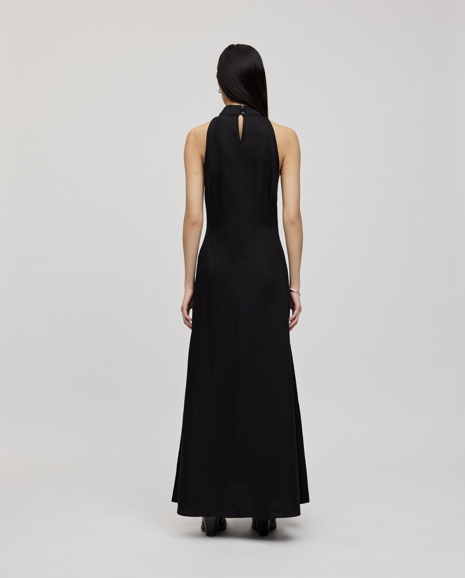 A woman is seen from the back against a plain light background, wearing the IVY OAK NELA Dress—a long, sleeveless black linen blend dress with an elegant neckholder design.
