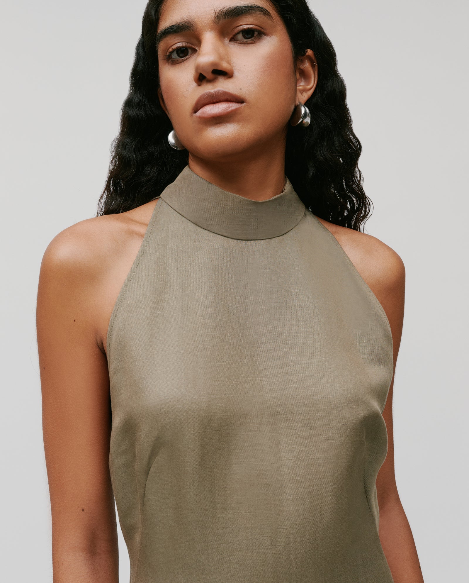 Woman wearing the IVY OAK NELA Dress, a sleeveless, high-neck, taupe maxi dress, styled with wavy hair and silver hoop earrings against a plain background.